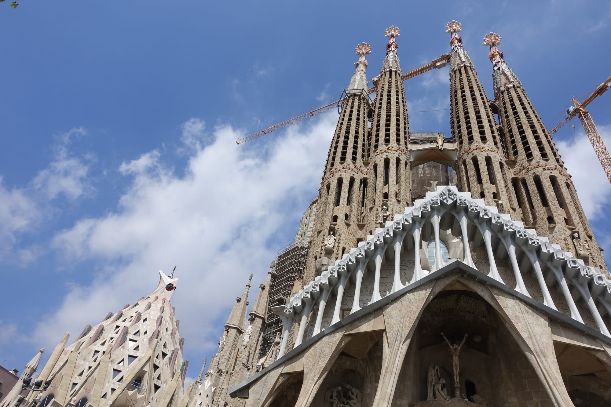 Four tall, perforated stone spires rise above a façade of angular, bone-like columns, with construction cranes swinging behind them against a blue sky
