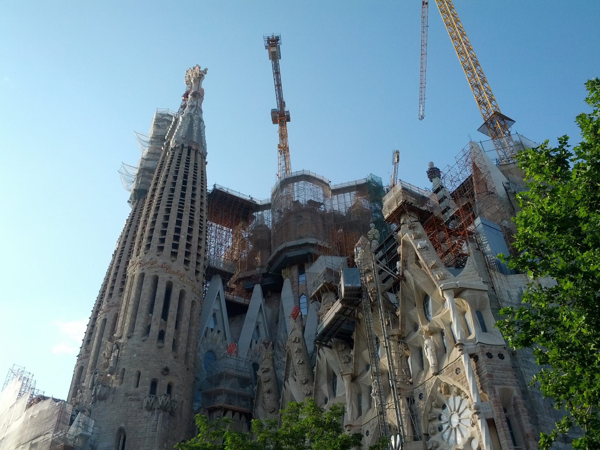 A tall, honeycombed stone spire beside unfinished towers wrapped in scaffolding, with two tower cranes rising above