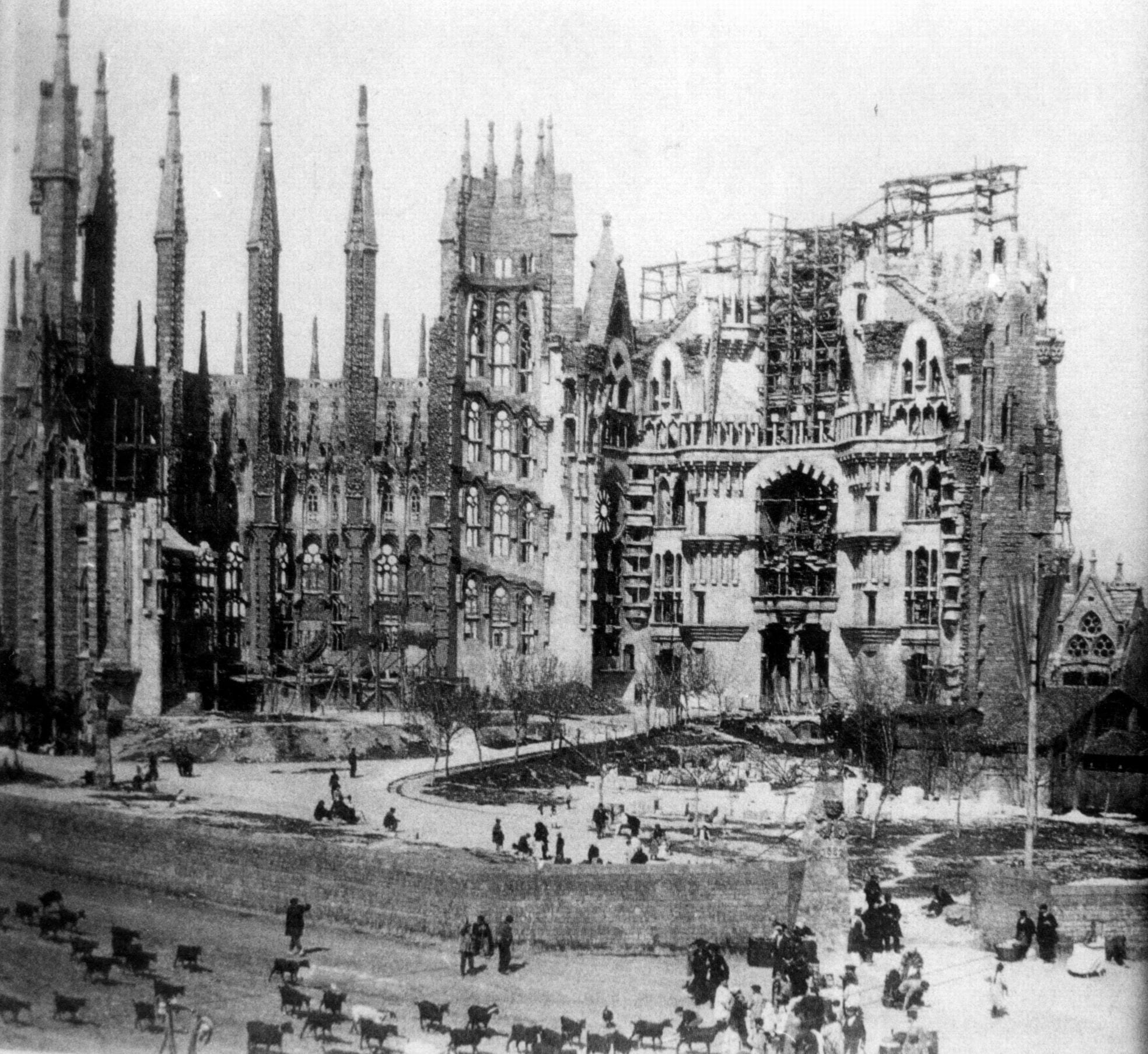 A black-and-white photograph of a half-built stone church with tall pointed spires on the left and scaffolding on the right, above an open dirt square with people and a flock of goats