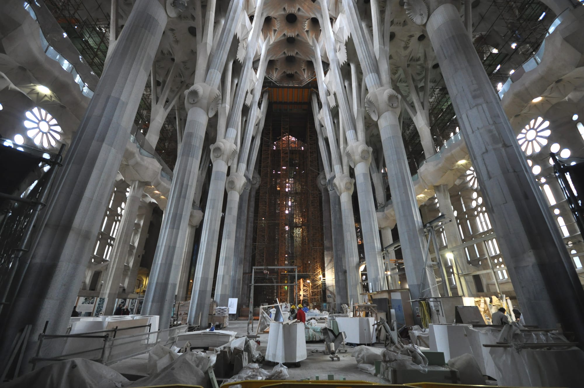Tall, pale tree-like columns branching into a star-patterned ceiling above a nave cluttered with building materials, scaffolding and workers in hard hats