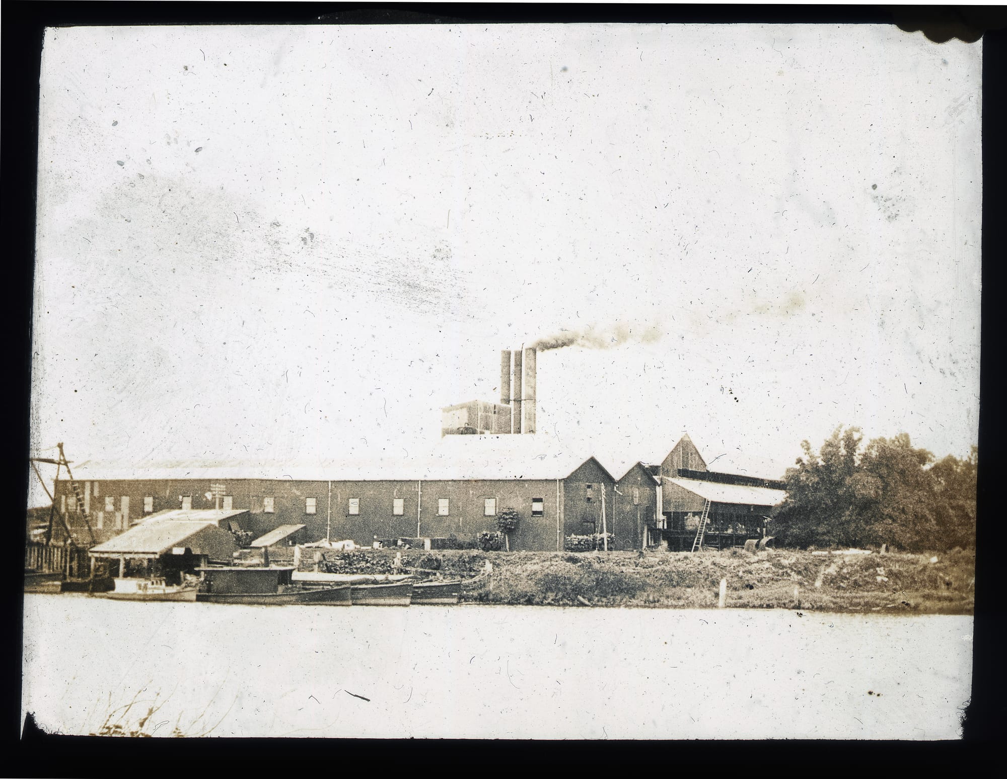An old sepia photograph of a long corrugated mill building with smoking chimneys beside a river with small boats