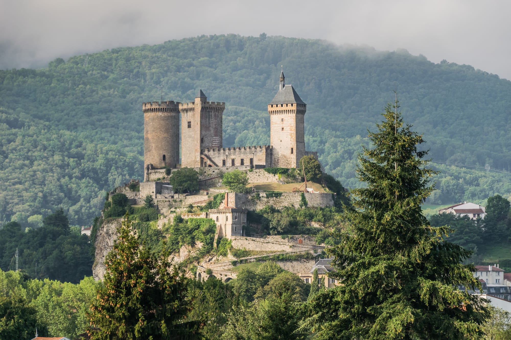A medieval castle with three tall stone towers on a rocky hilltop above a town, with a wooded hill behind