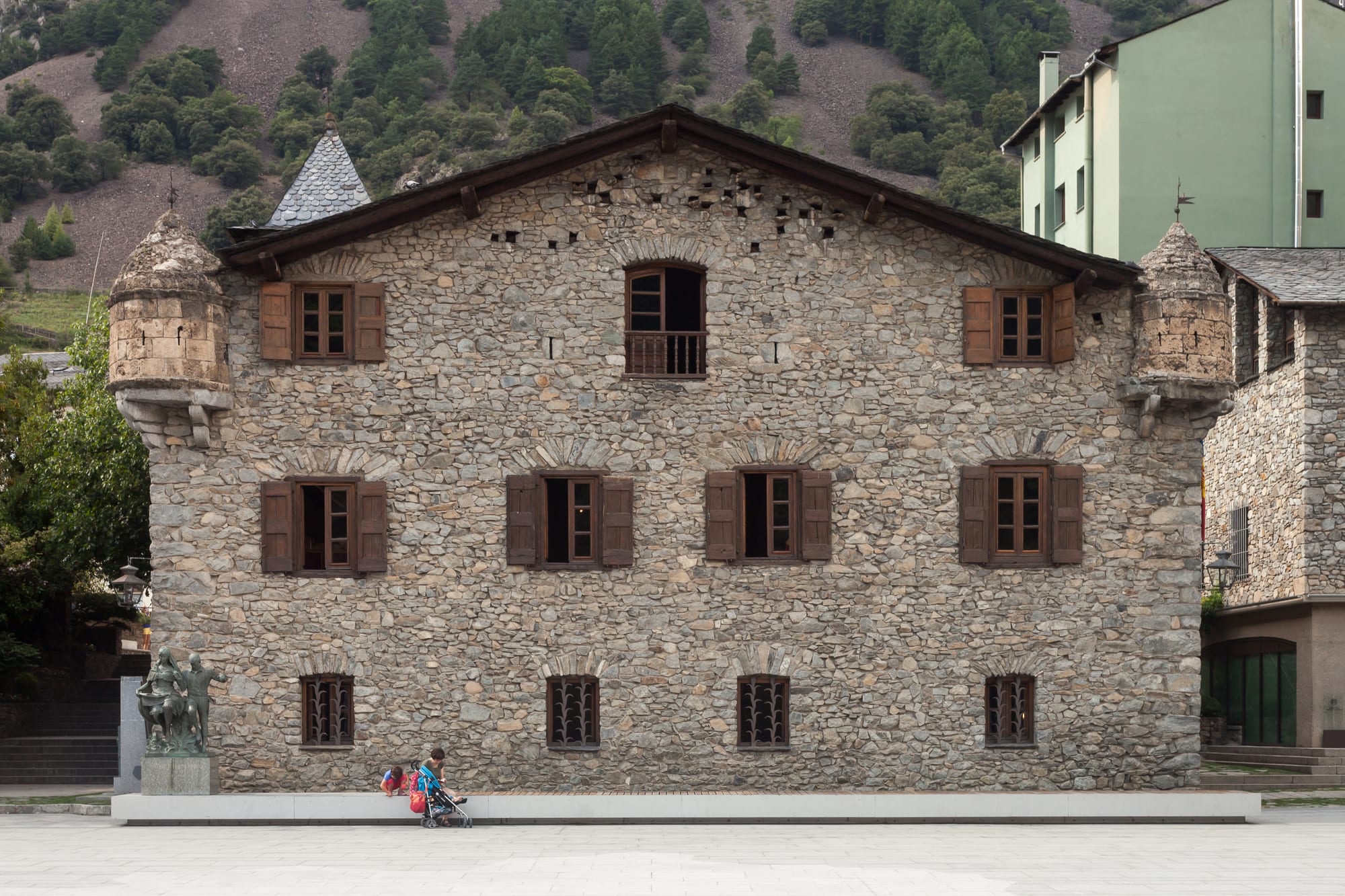 A three-storey stone house with wooden shutters and small corner turrets on a paved square, with a wooded mountainside behind