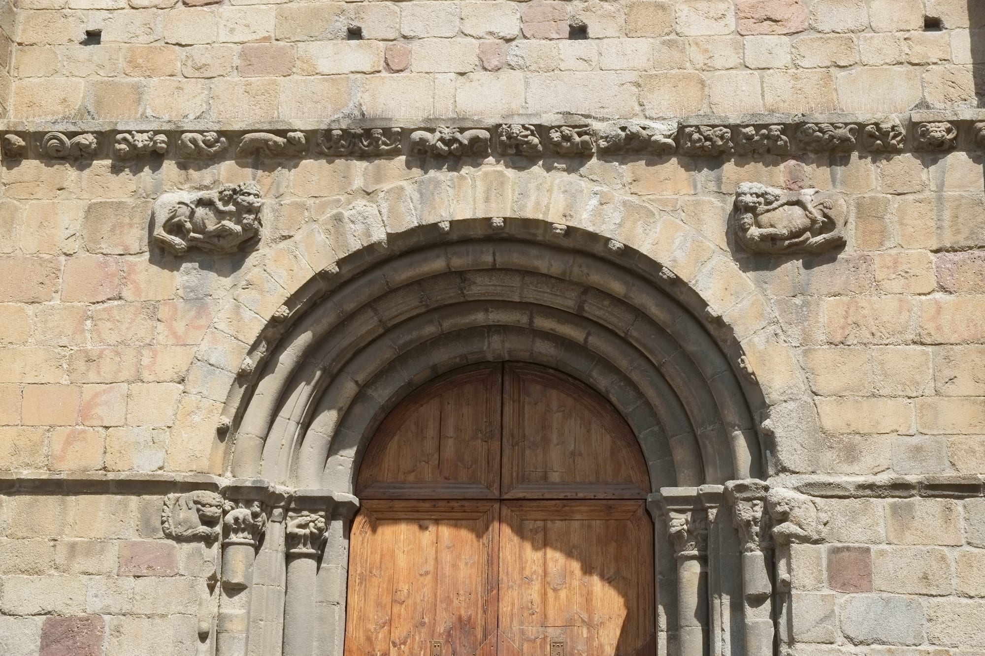 A round-arched Romanesque stone doorway with wooden doors, beneath a carved frieze of small animal and human figures