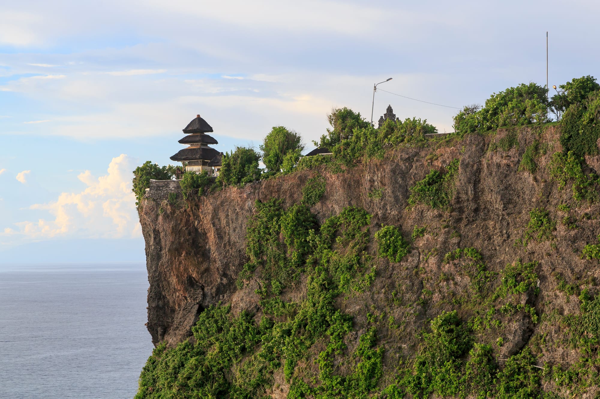 A small temple with tiered black thatched roofs on the edge of a high cliff above the sea