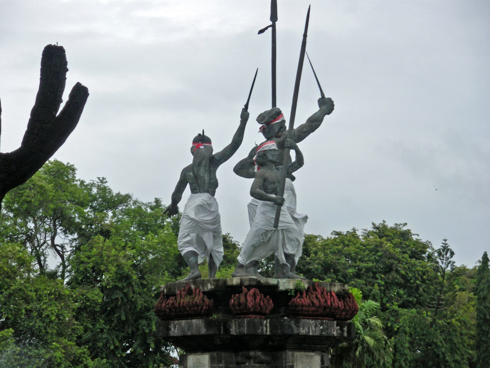 Bronze statues of a man, a woman and a child holding spears and a dagger aloft, on a stone plinth among trees