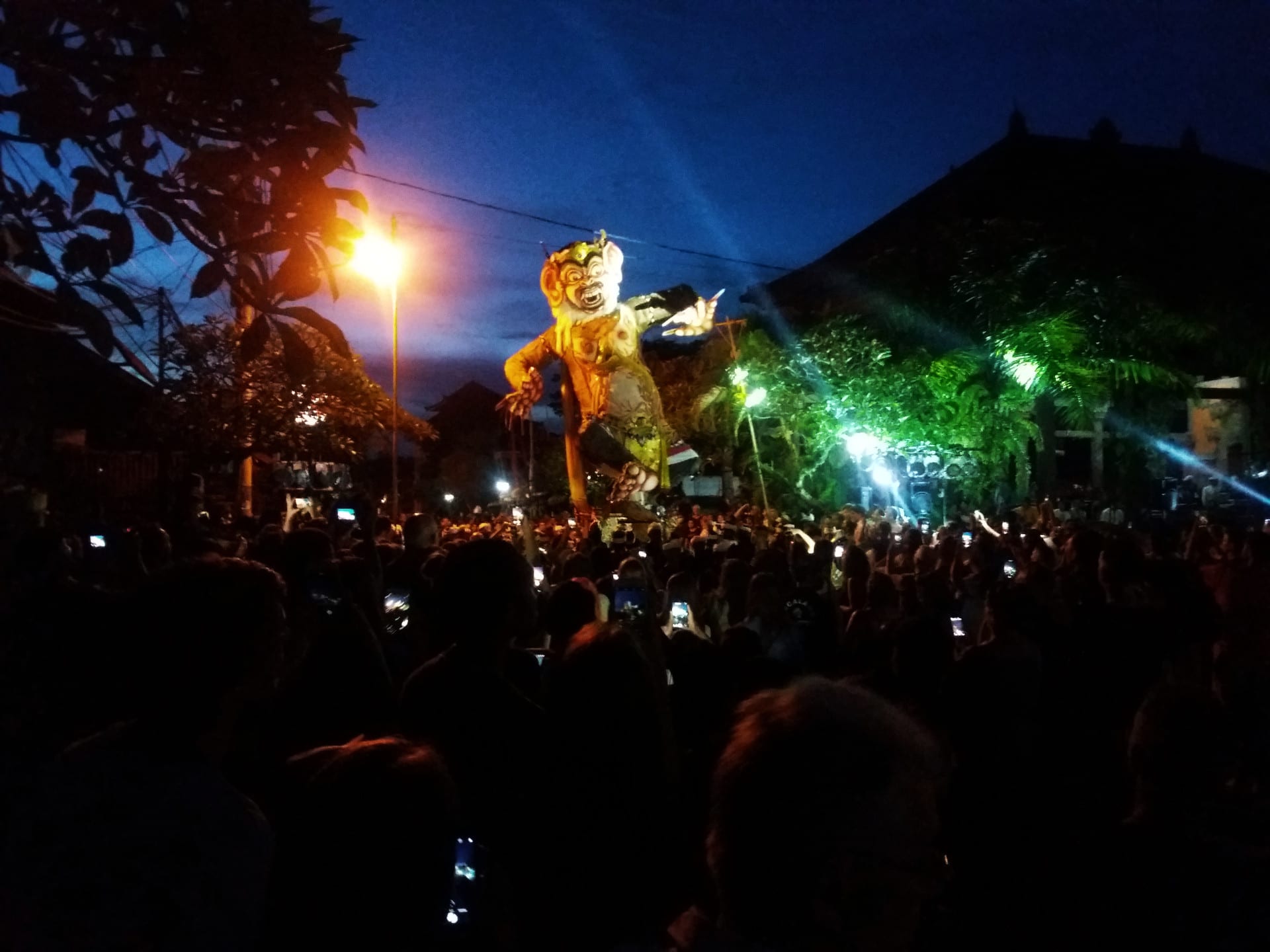 A giant illuminated demon figure raised above a dense crowd at night, lit by streetlights