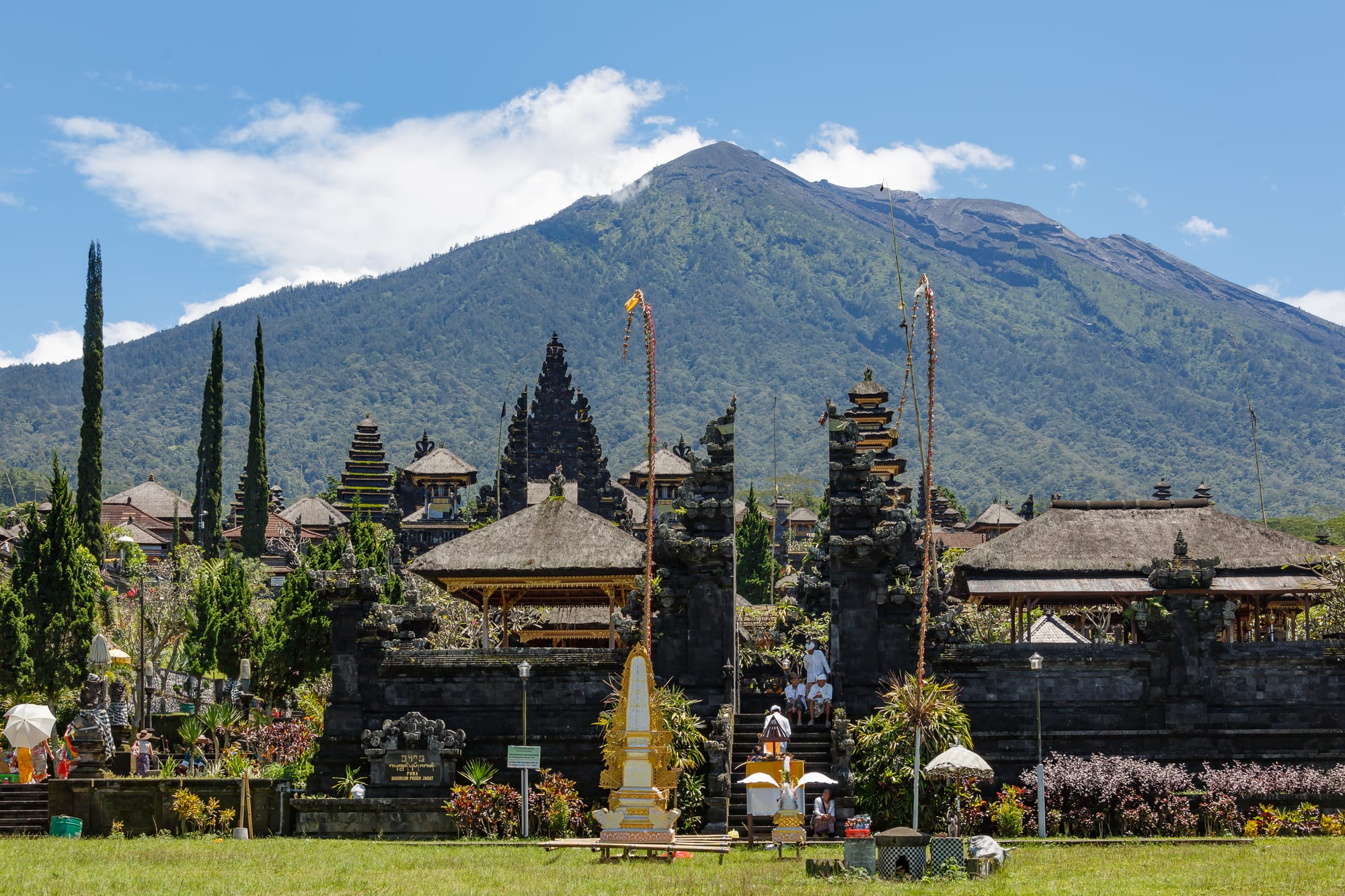 Black tiered shrine towers and thatched pavilions of a temple complex below a large green volcano