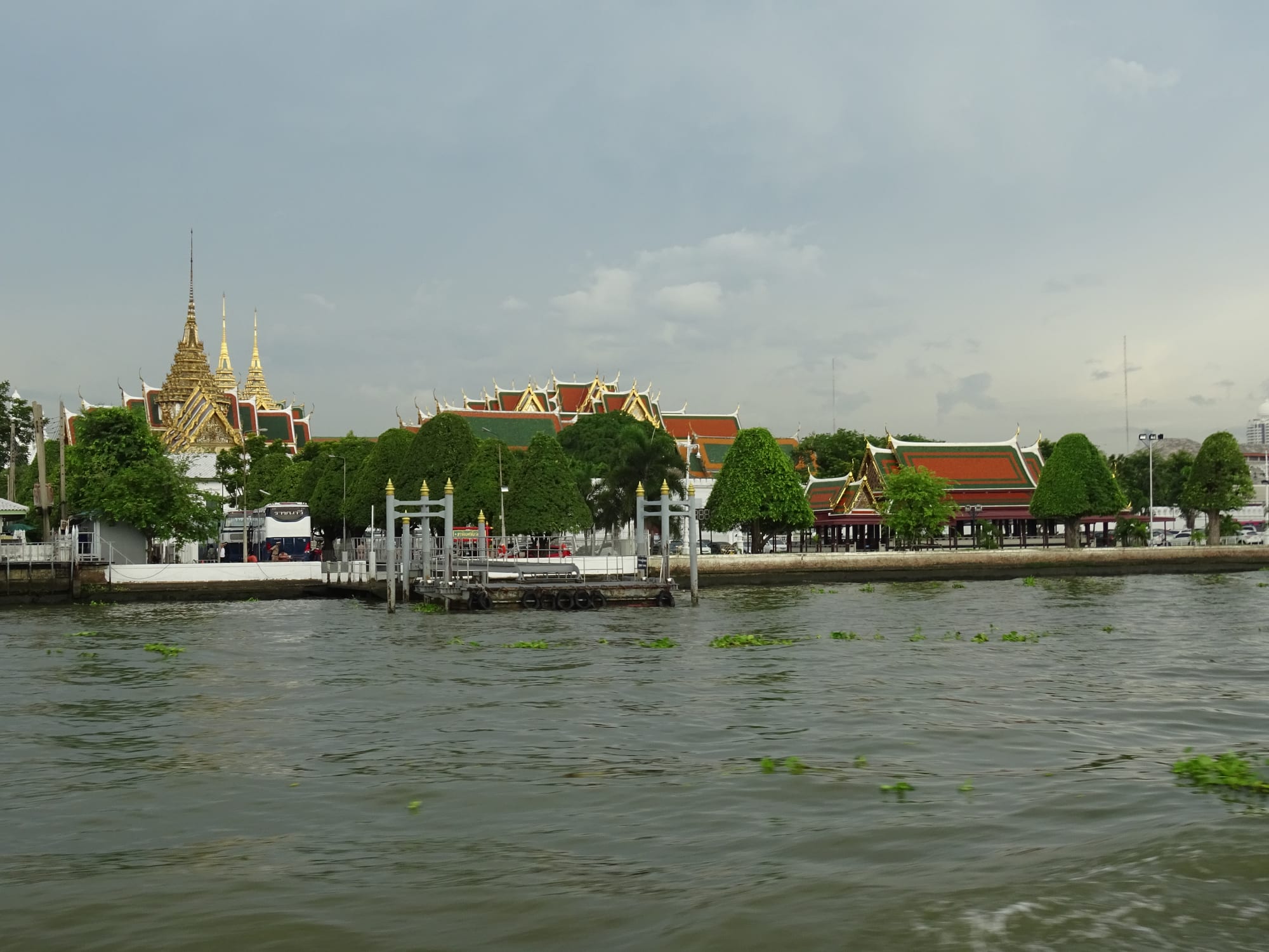 Gilded spires and red-and-green tiled roofs of a palace complex among trees on the bank of a wide brown river