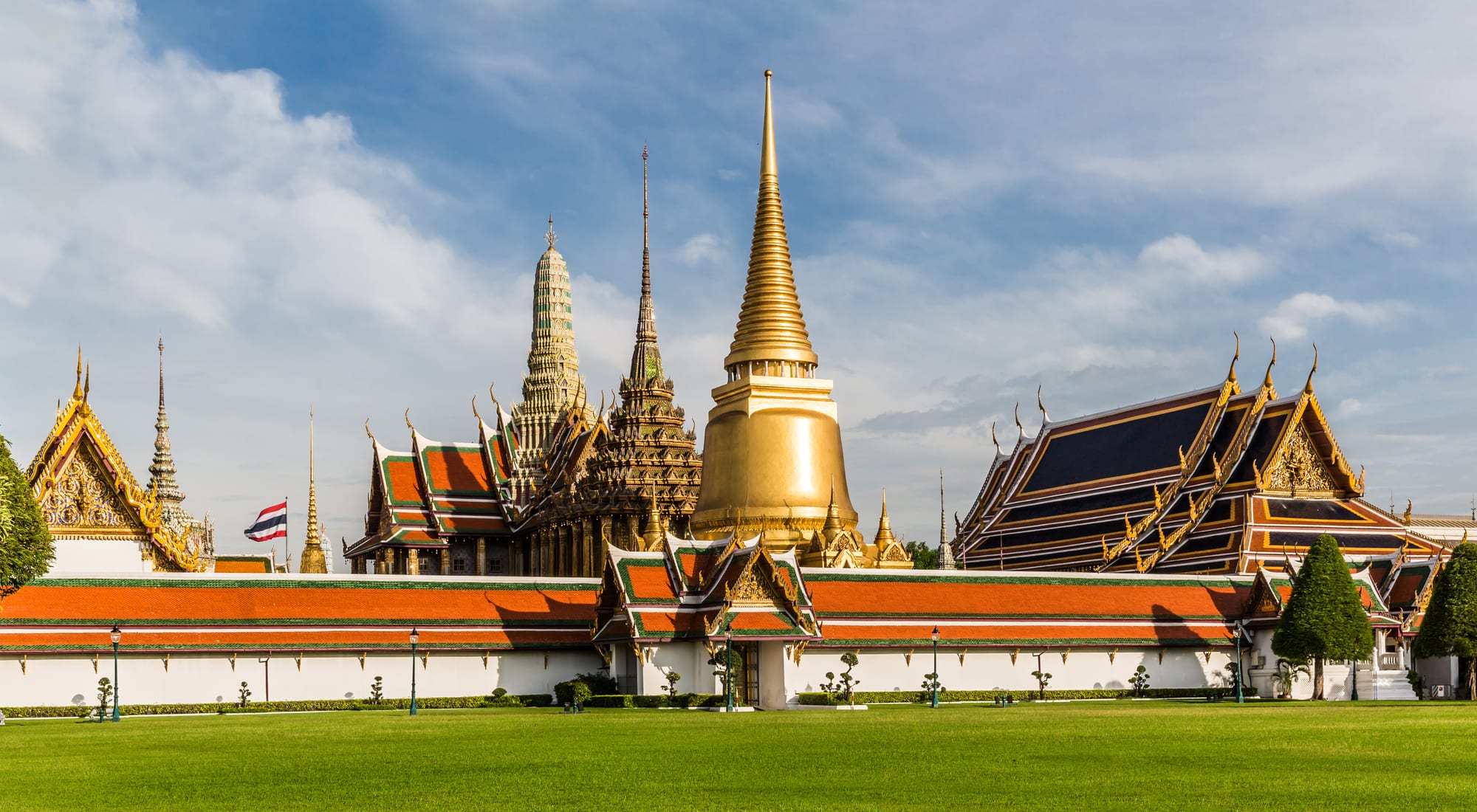 A golden bell-shaped stupa, a tall spired pavilion and orange-and-green tiled temple roofs behind a white wall and a green lawn