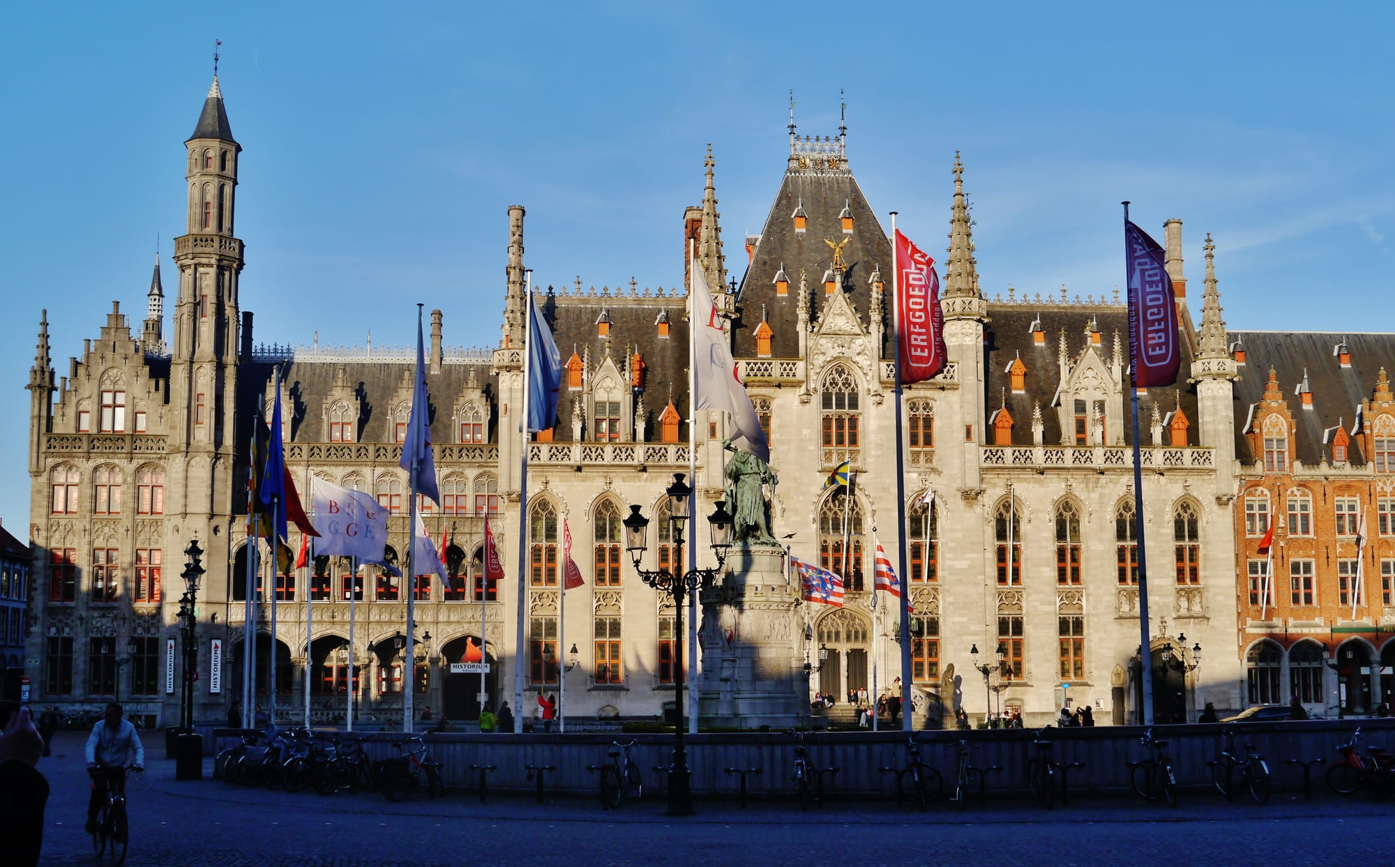 An ornate pale stone building with pointed windows, towers and spires behind a statue and a row of flags on a large square