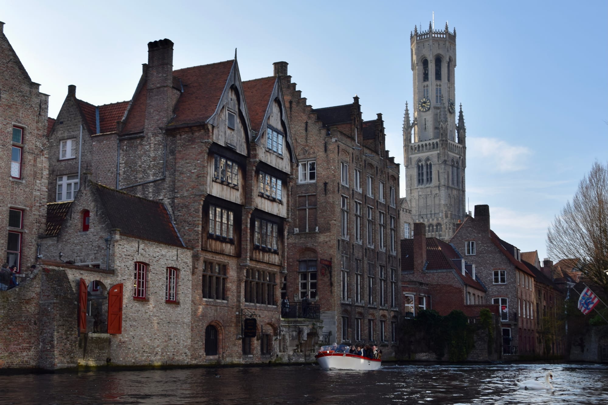 Old brick houses with stepped and pointed gables rising straight from a canal, a tour boat on the water and a tall stone bell tower behind