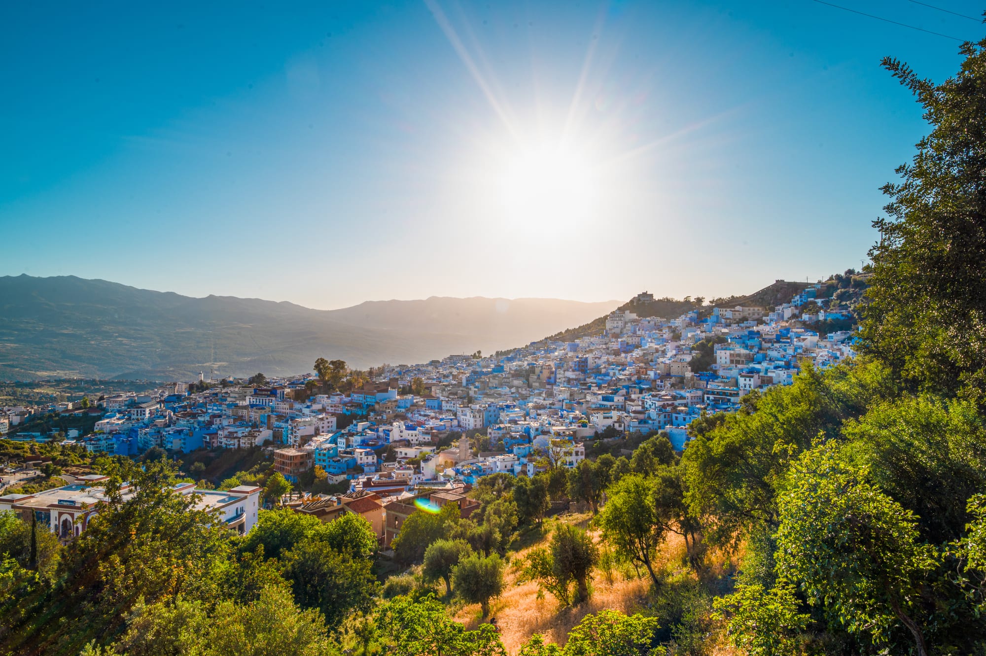 A hillside town of white and blue houses spread across a slope, with forested mountains behind under a low evening sun