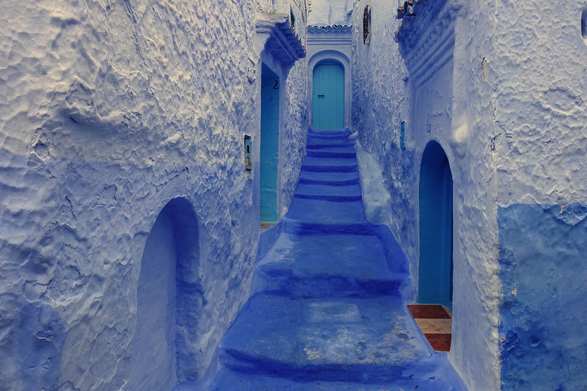 A narrow stepped alley between thick whitewashed walls washed in shades of blue, leading up to a turquoise door under an arch