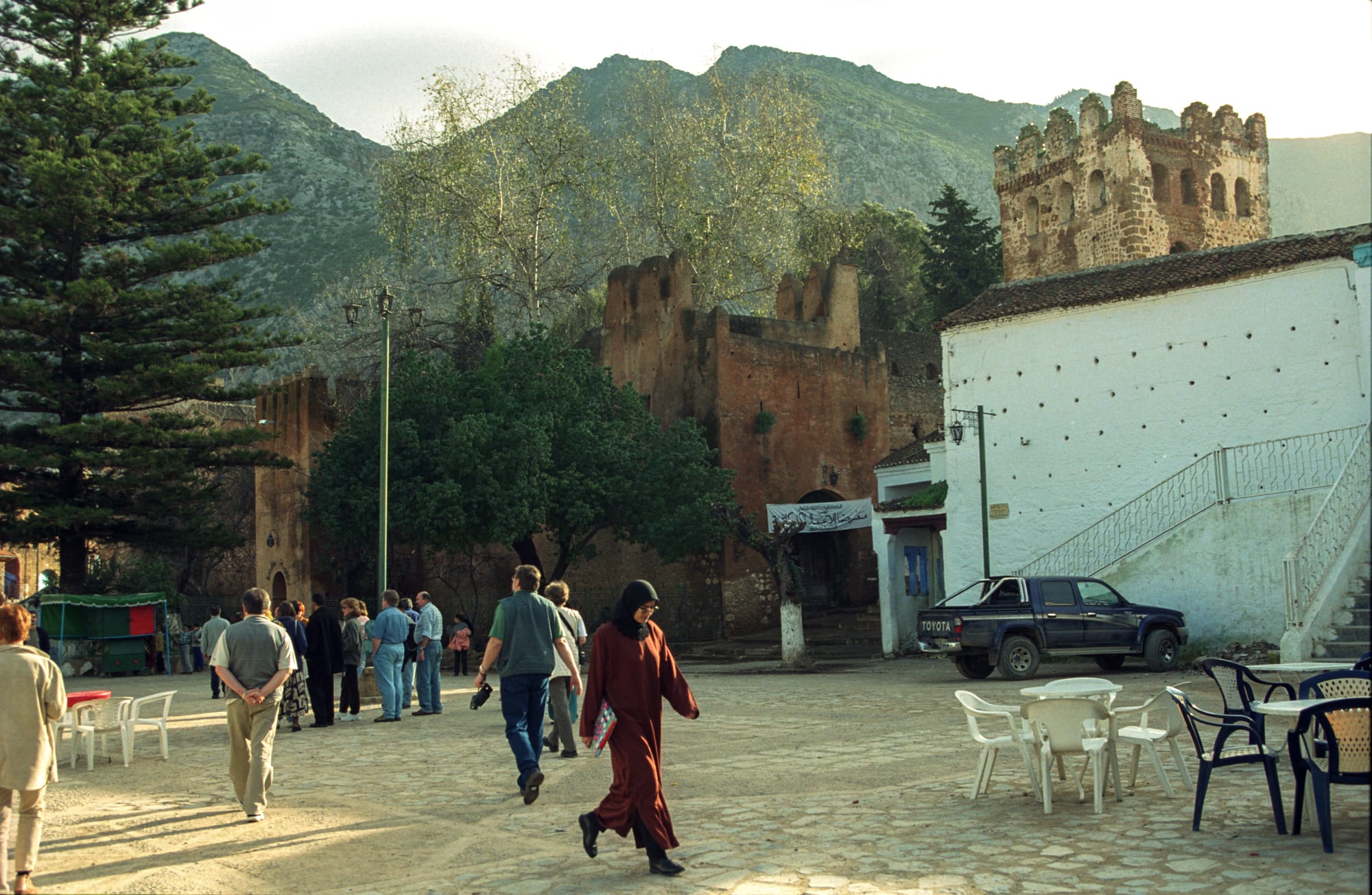 A cobbled town square with café tables, people walking, a reddish-brown crenellated fortress and a white mosque, with mountains rising behind