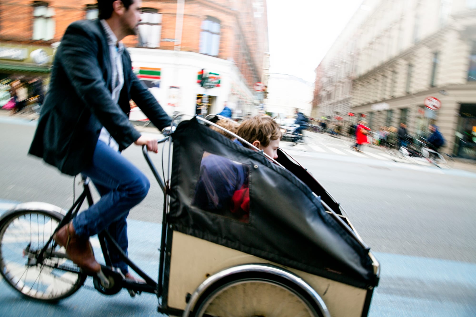 A man in a blazer pedals a three-wheeled cargo bike with two small children sitting in a covered wooden box at the front