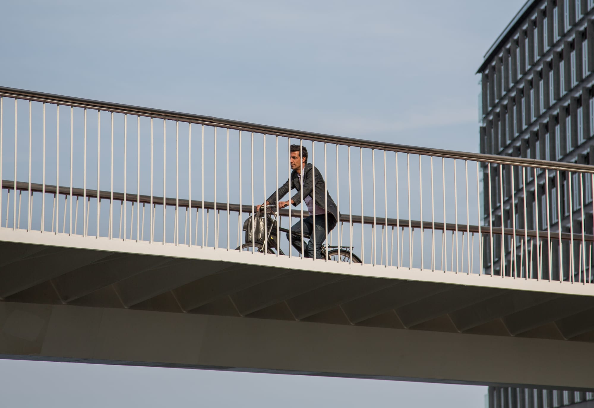 A man in a jacket rides a bicycle across a white elevated bridge with thin railings, against a pale sky