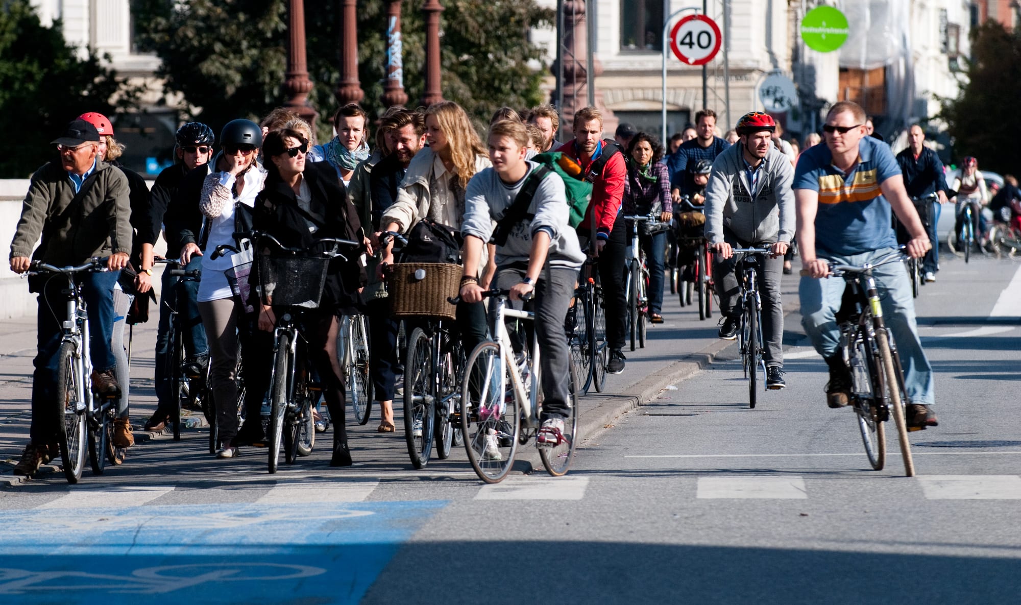 A crowd of cyclists in everyday clothes, some with baskets and a few with helmets, gathered at a crossing on a city street on a sunny day
