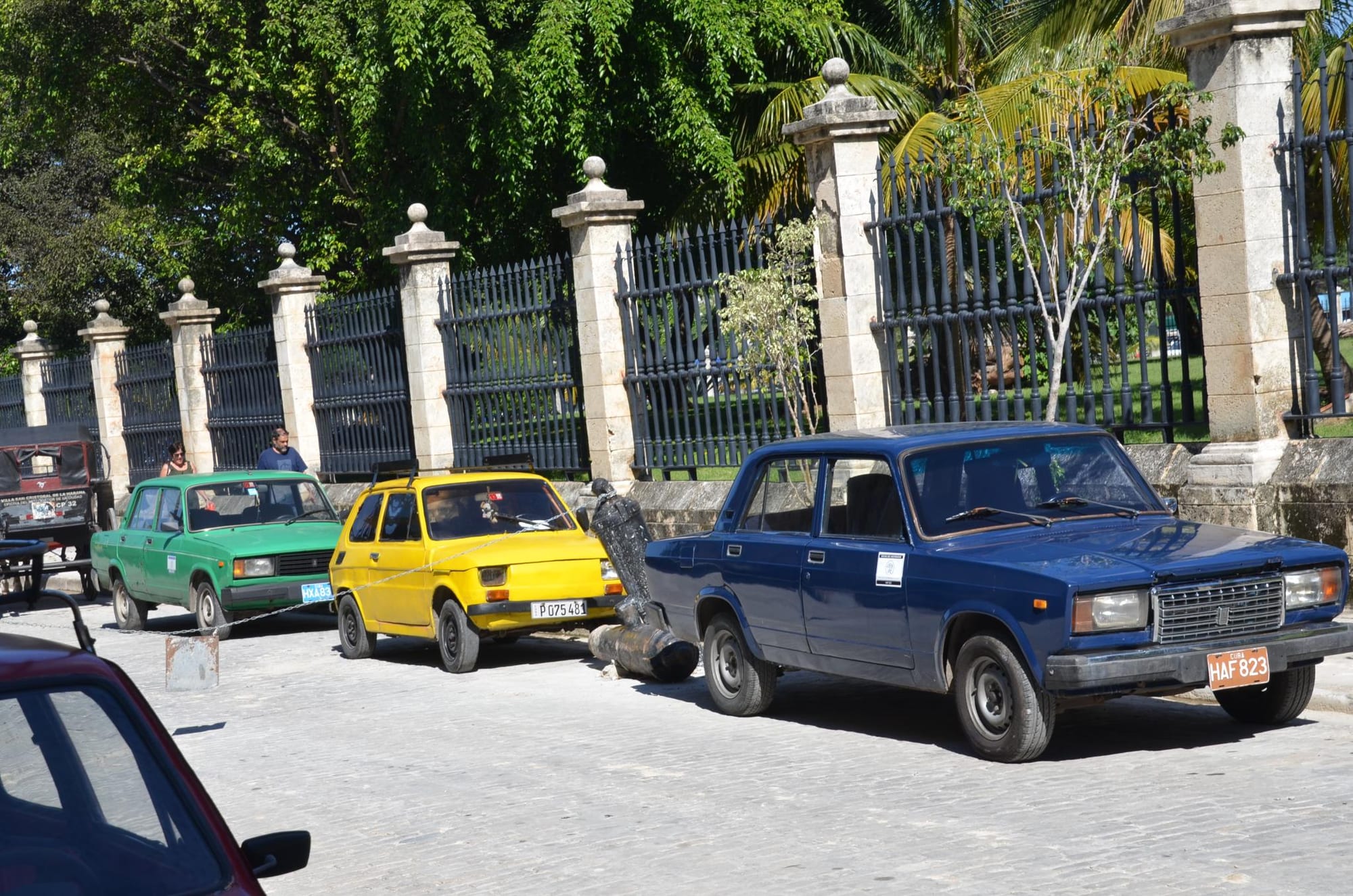 A green and a blue boxy Soviet-era Lada saloon and a small yellow Fiat parked on a cobbled street beside an iron fence