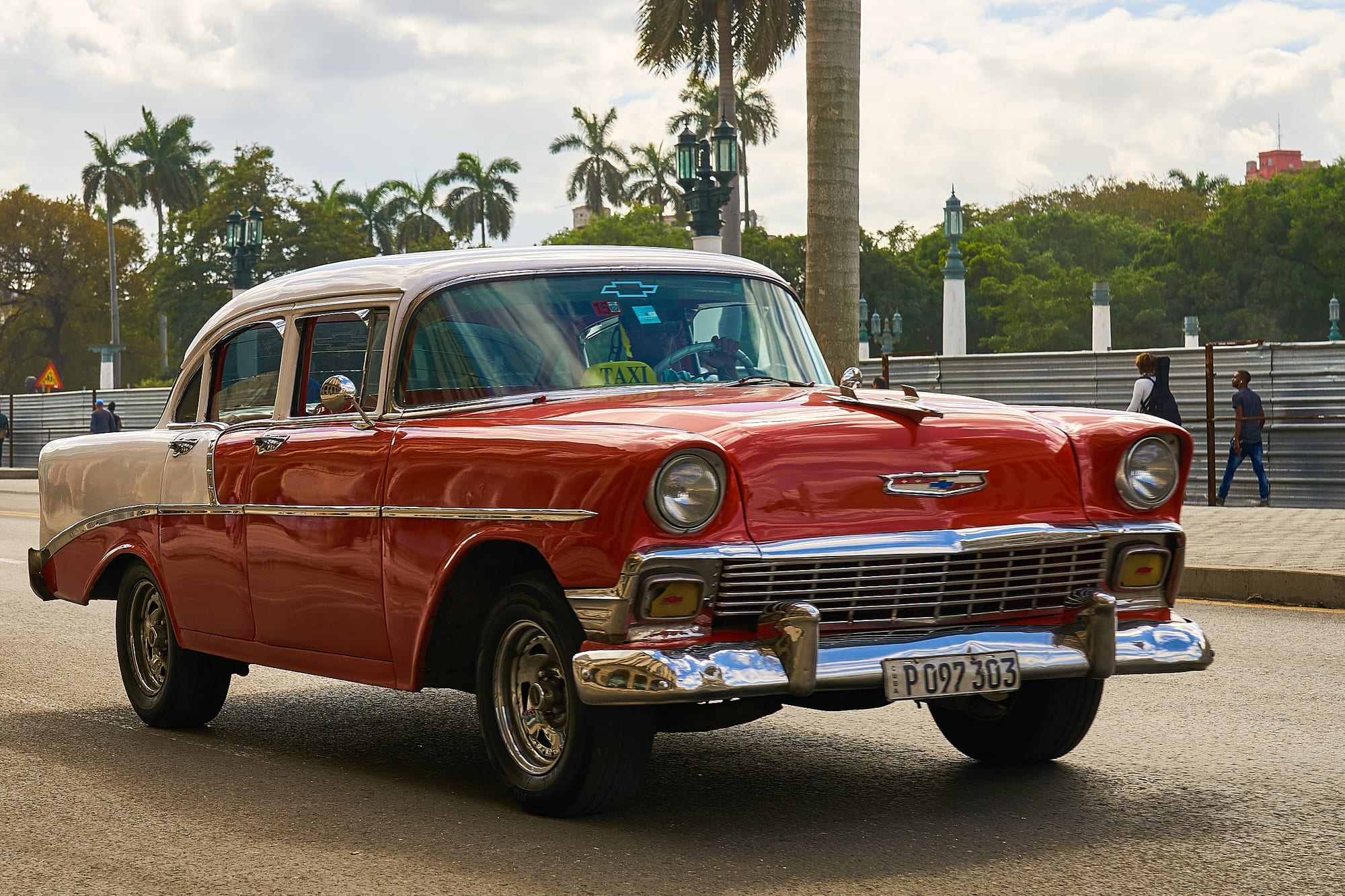 A red and white 1950s American sedan with chrome bumpers and a taxi sign, driving along a palm-lined street