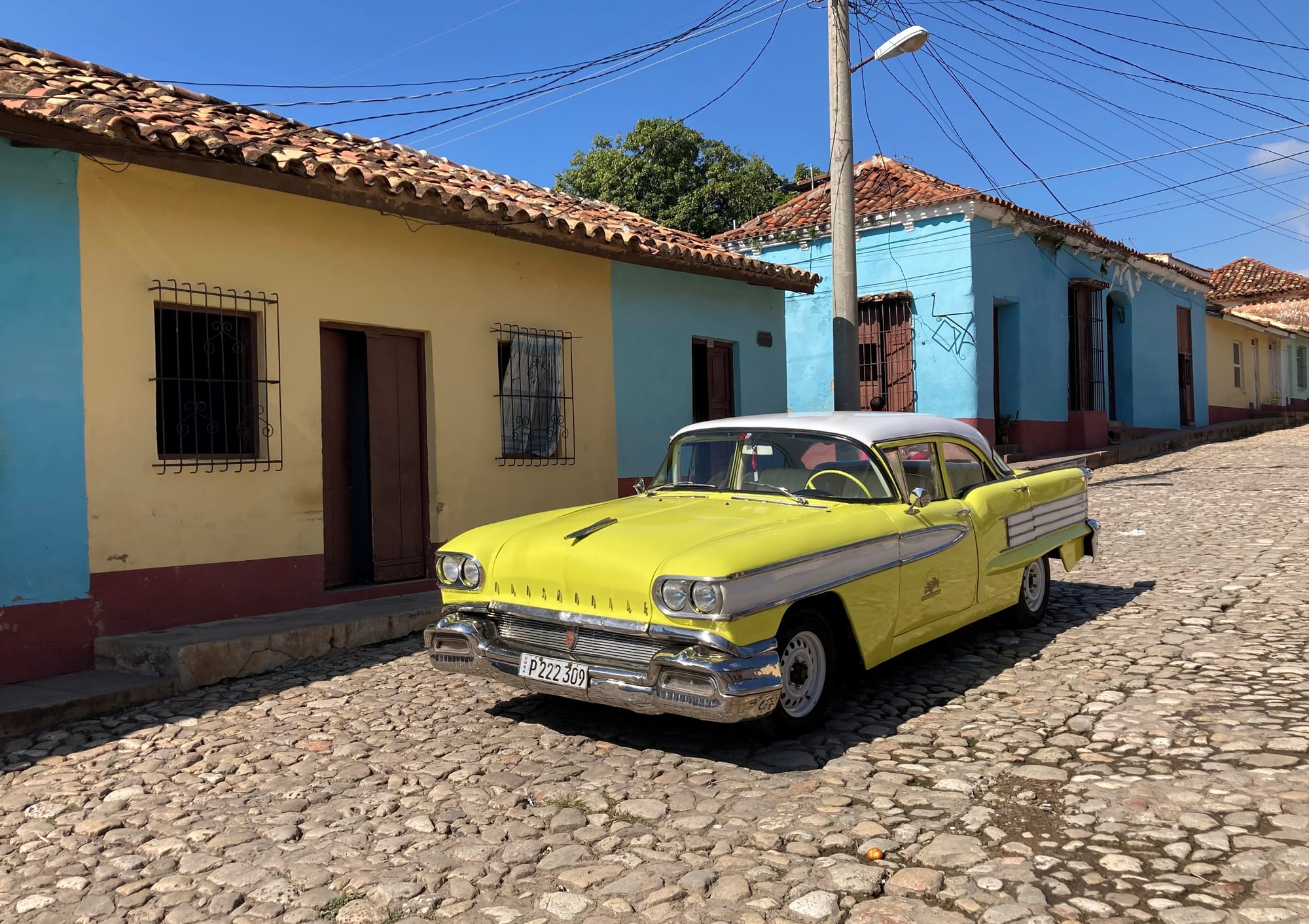 A yellow and white 1950s American saloon parked on a cobbled street lined with low yellow and blue colonial houses