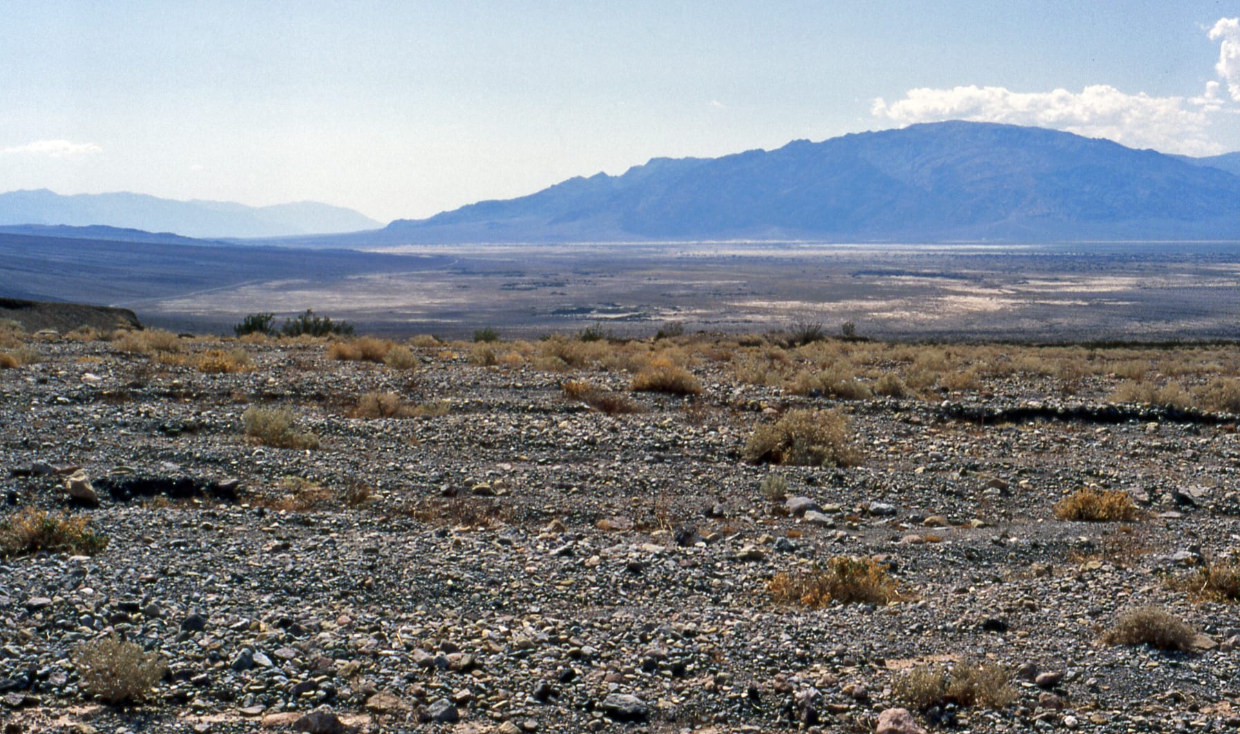 A stony slope of gravel and small desert shrubs leading down to a wide, pale valley floor, with a bare mountain range under a hazy sky