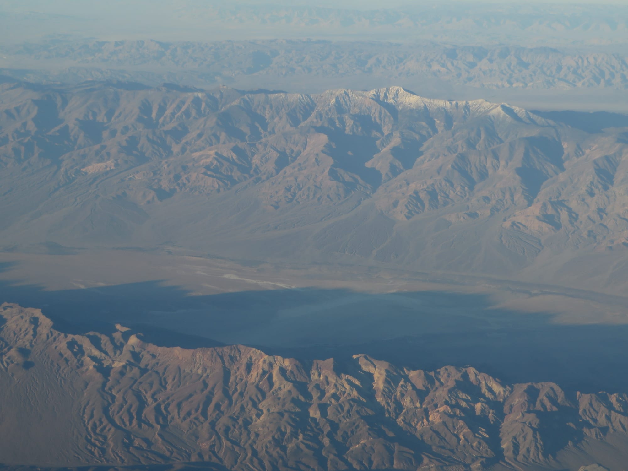 An aerial view of a long, flat, pale valley floor between bare brown mountain ranges, with a snow-dusted summit beyond