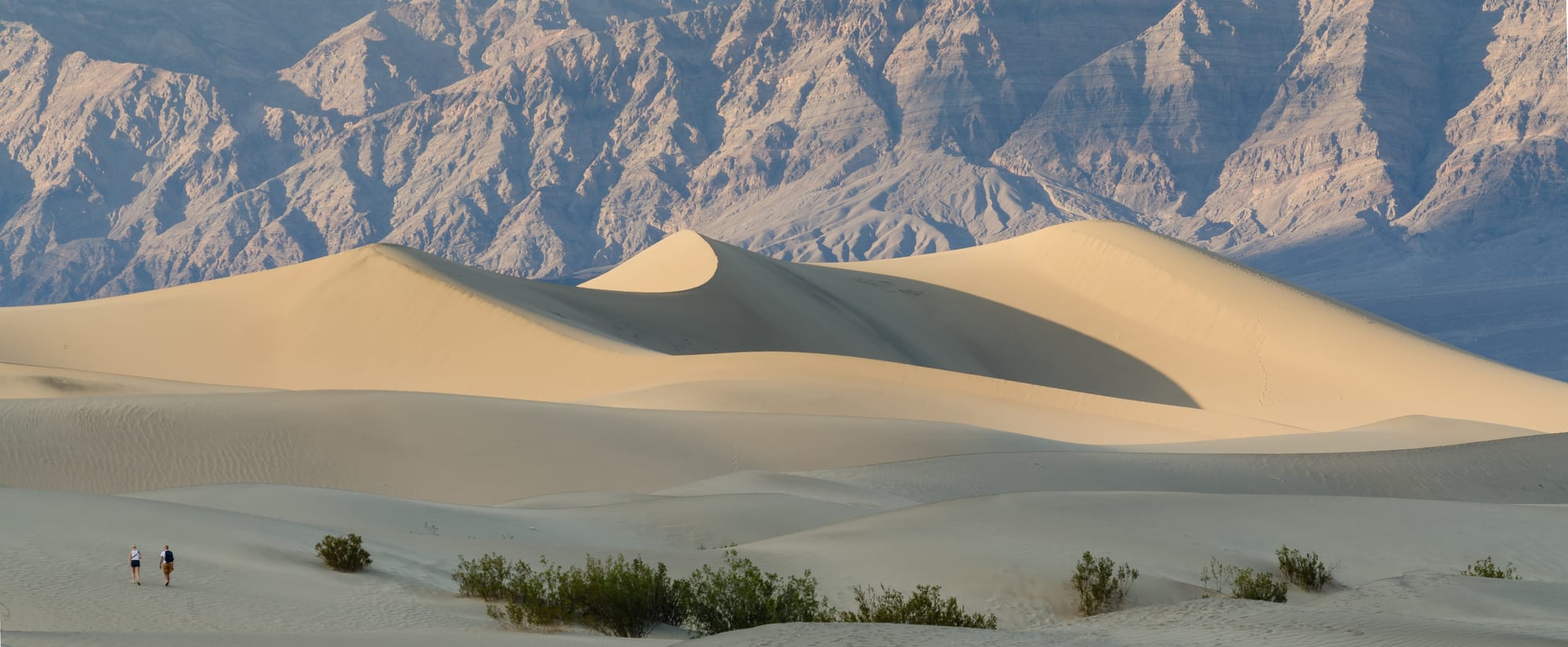 Smooth, pale sand dunes dotted with small green shrubs, two walkers at the edge, beneath a steep, bare, rocky mountain wall
