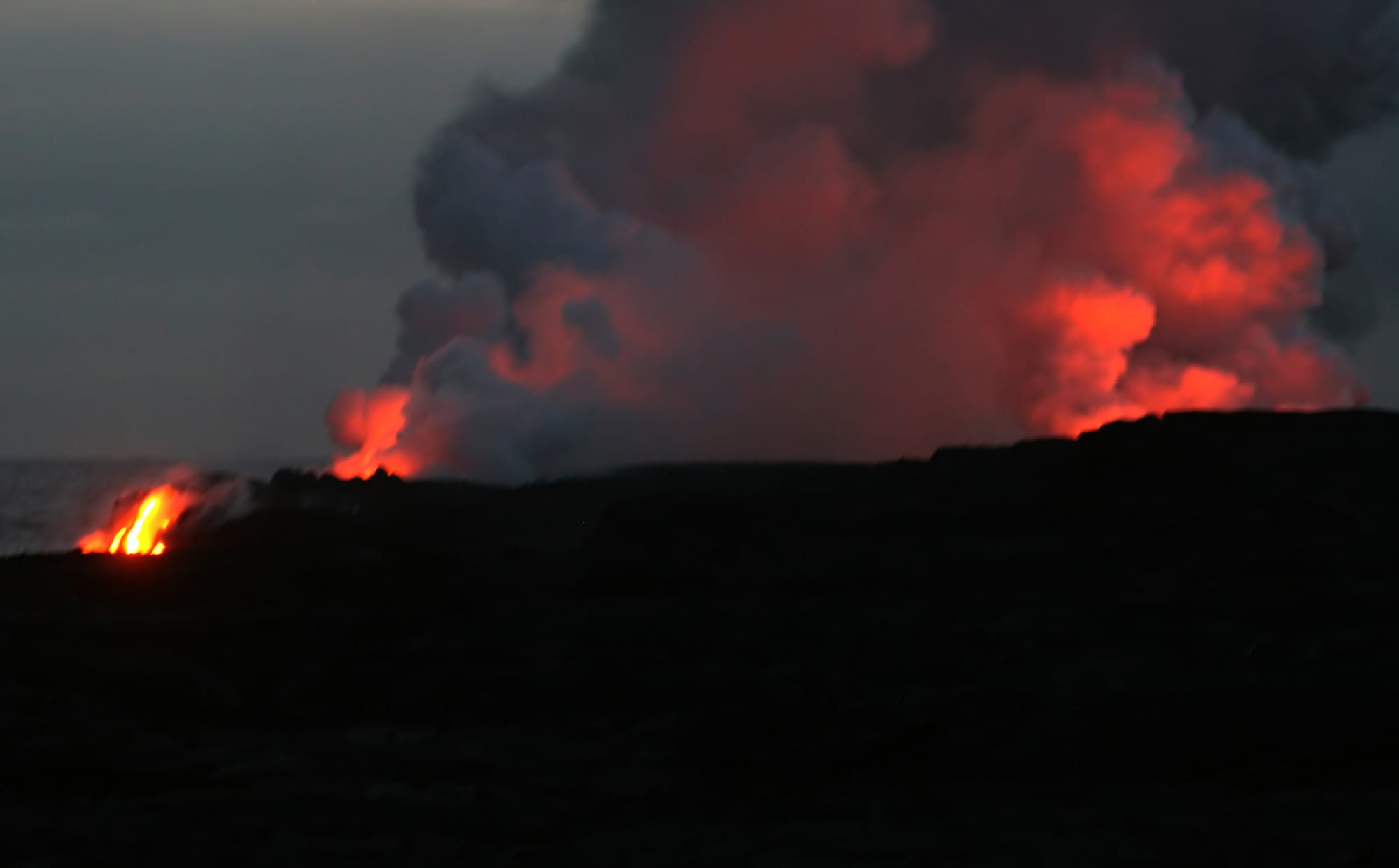 At dusk, glowing orange lava pours into the sea at the edge of a dark lava shore, beneath a towering plume of red-lit steam