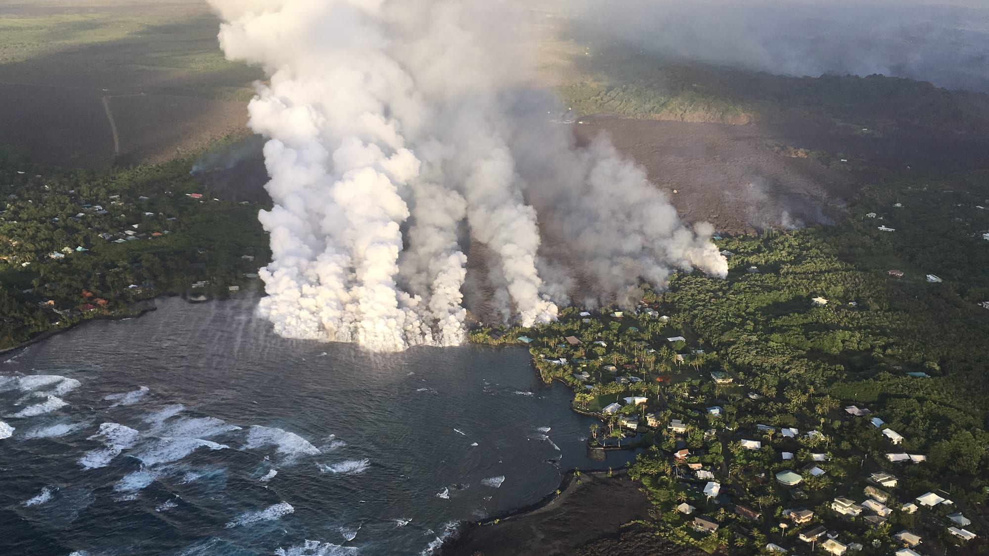 An aerial view of huge white plumes of steam rising where a dark lava flow meets the sea beside a coastal neighbourhood of houses among trees