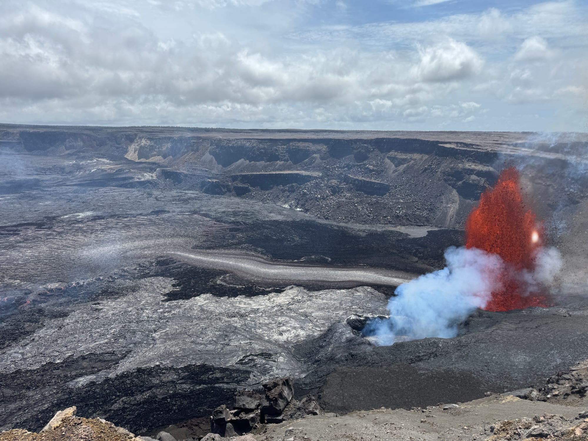 A wide, grey crater floor of fresh lava with a red fountain of molten rock and white steam erupting at its edge