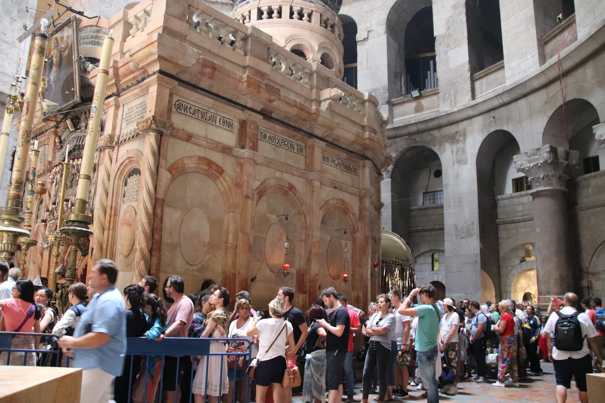 A crowd of visitors queuing beside an ornate pinkish stone shrine with candlesticks, beneath a tall rotunda of arches