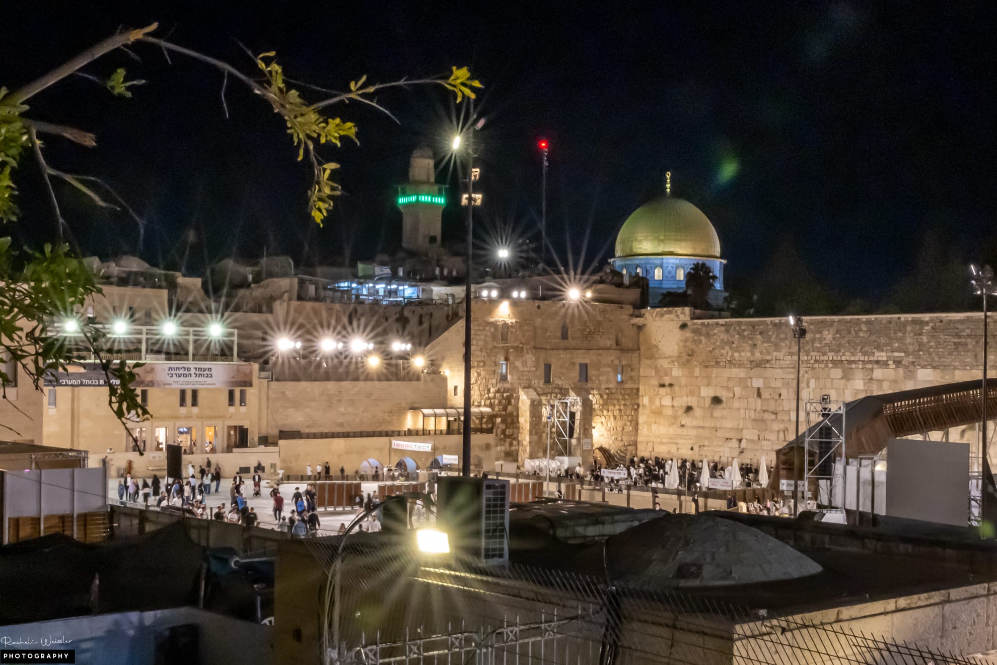 Worshippers in a floodlit plaza at night before a wall of huge pale stone blocks, with a golden dome rising beyond the top of the wall