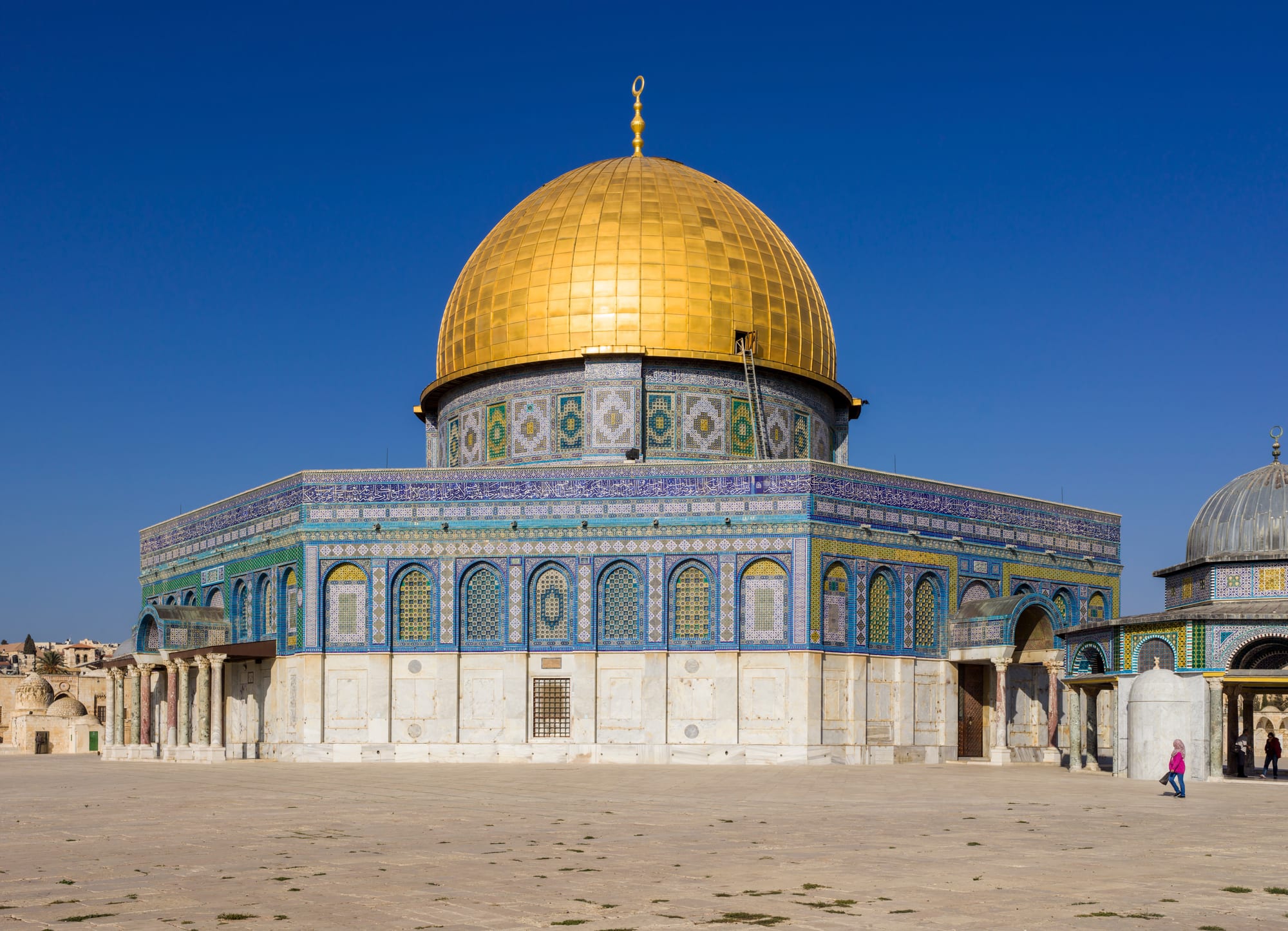 An octagonal shrine faced with blue, green and white tiles, topped by a large gleaming gold dome, standing on a wide stone plaza