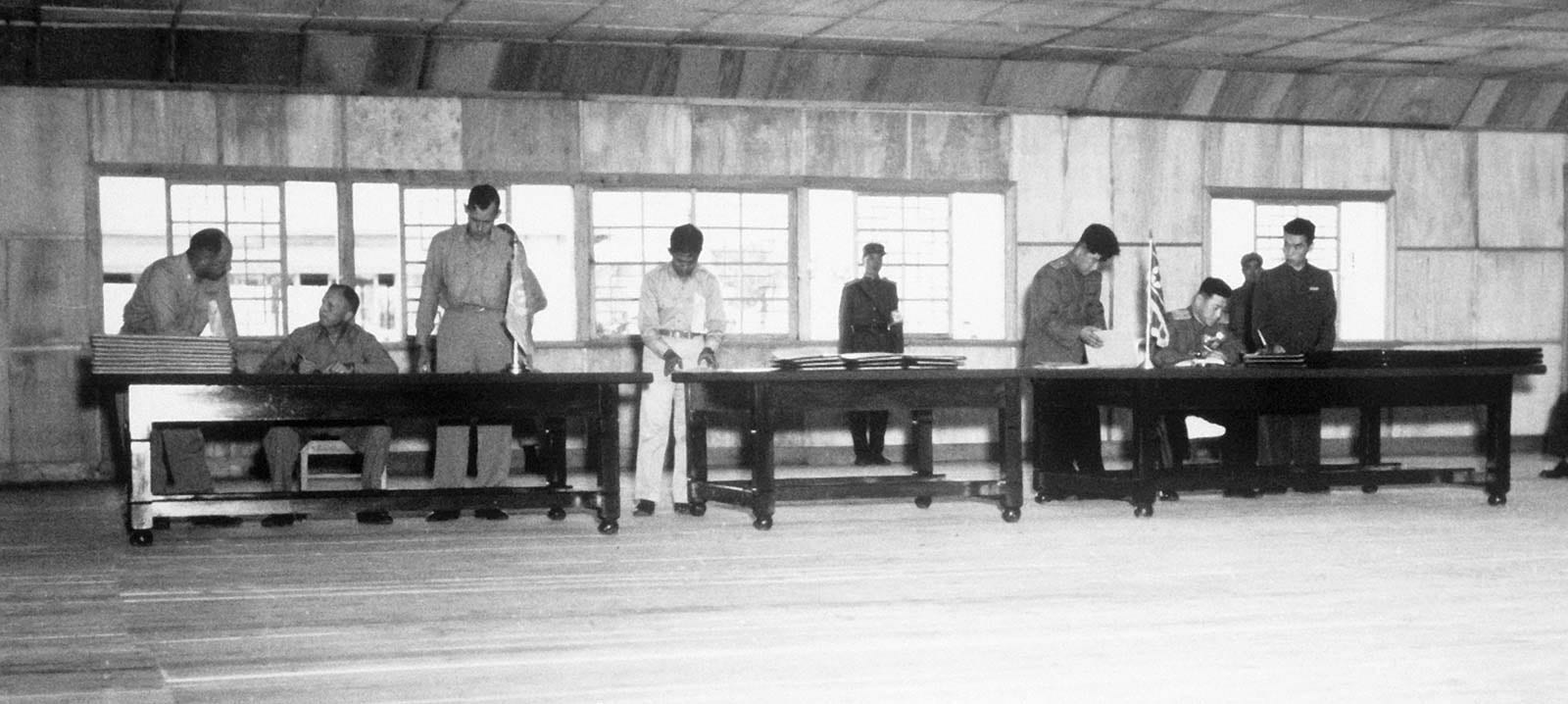 A black-and-white photograph of uniformed officers seated at long wooden tables in a plain wooden hall, signing documents, with flags on the tables