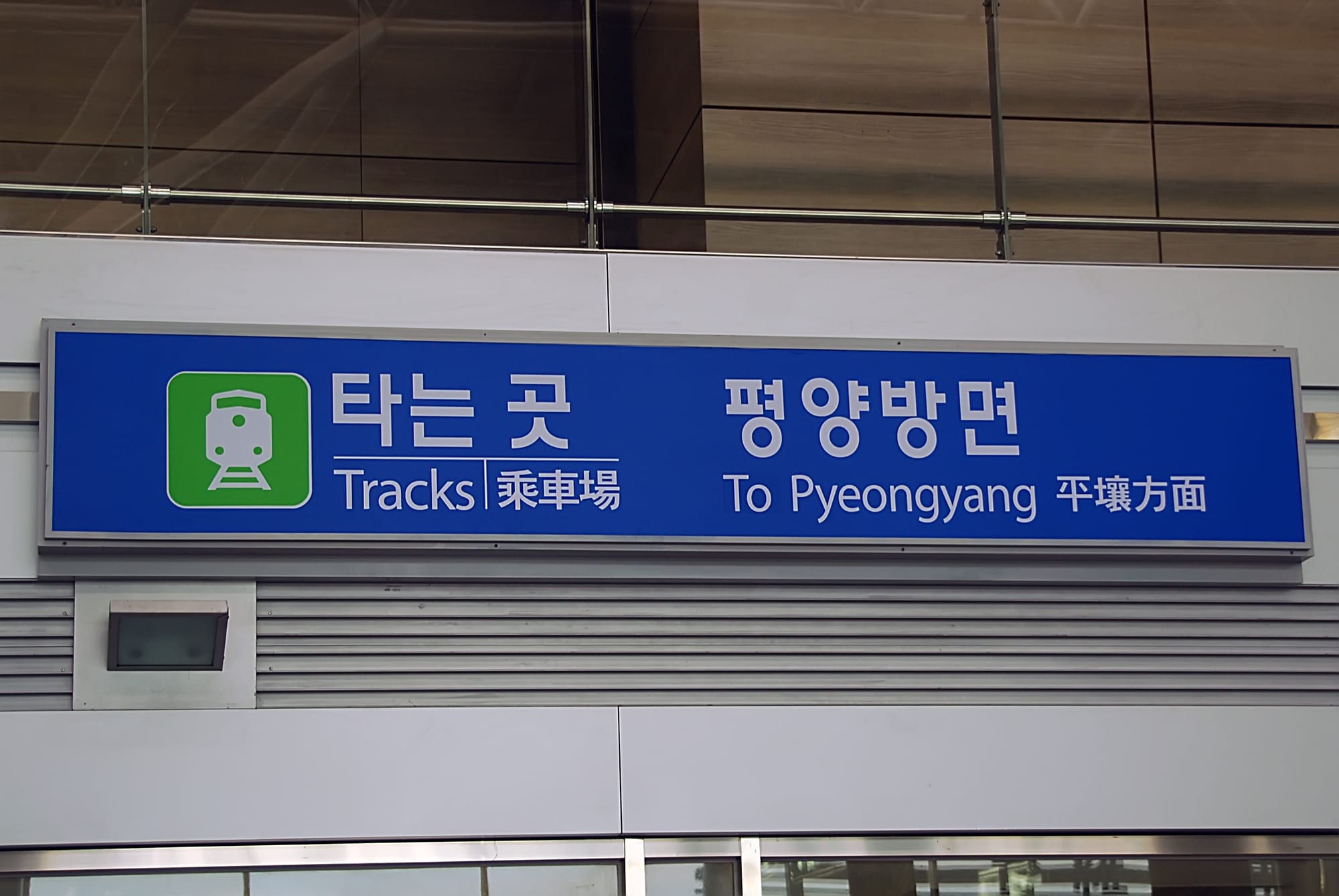 A blue station sign with a green train symbol reading Tracks and To Pyeongyang in Korean, English and Chinese