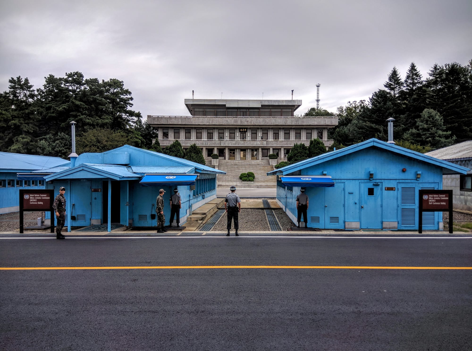 Two pale blue single-storey huts on either side of a gravel strip with uniformed guards standing between them, and a large grey stone building on the far side
