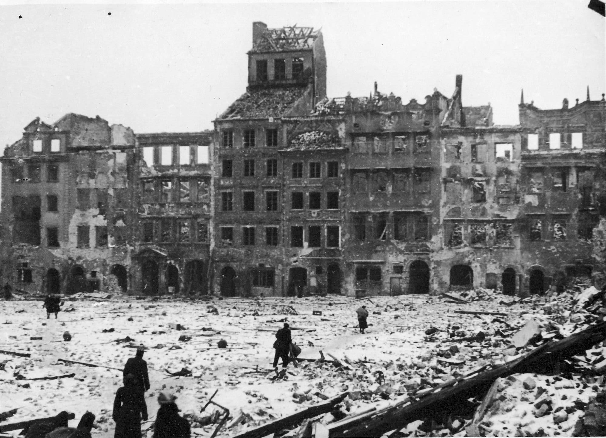 A black and white photograph of burnt-out, roofless townhouses around a snow-covered square strewn with rubble, with a few people walking through