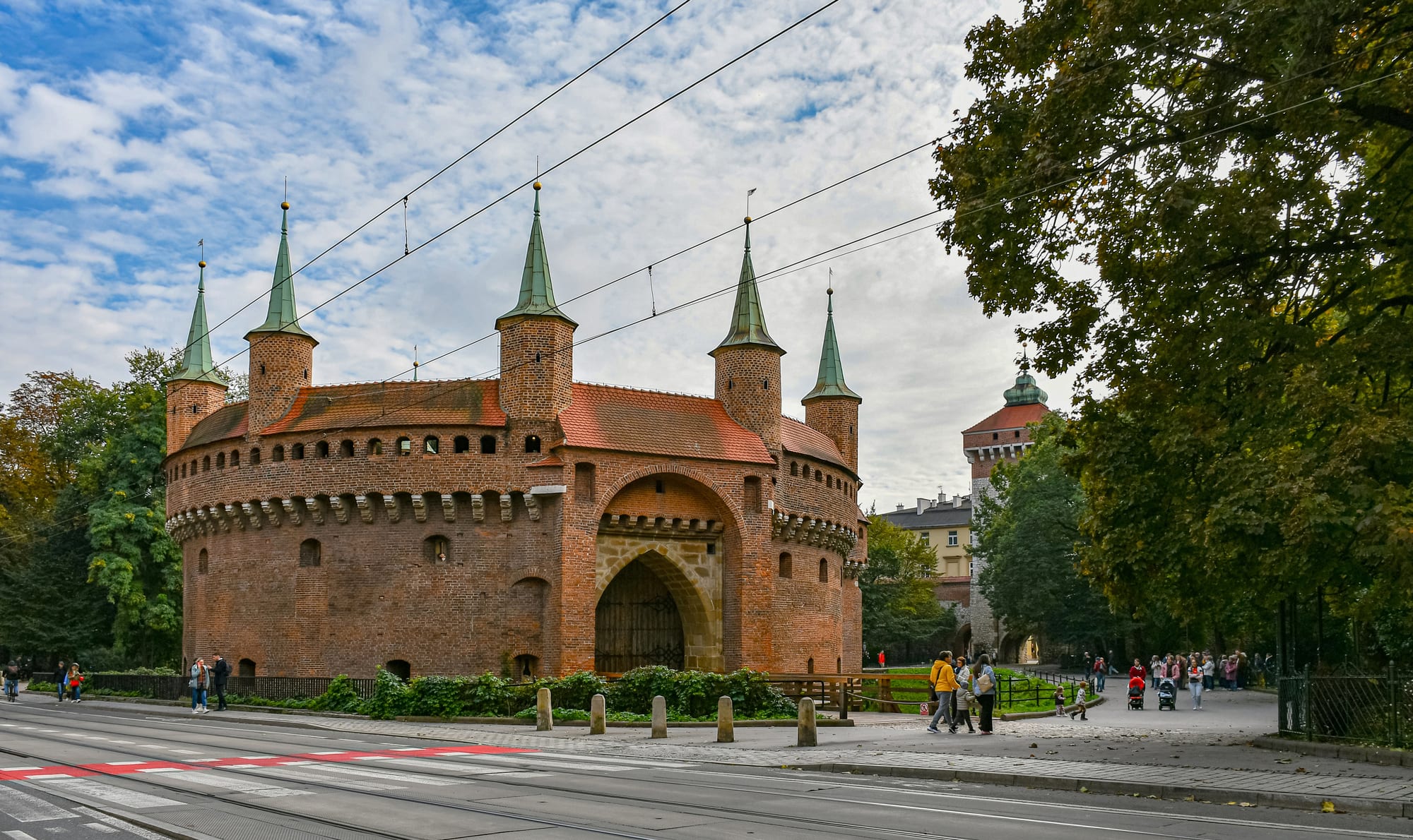 A round red-brick fortification with small pointed towers and a gateway, with a taller gate tower behind it among autumn trees