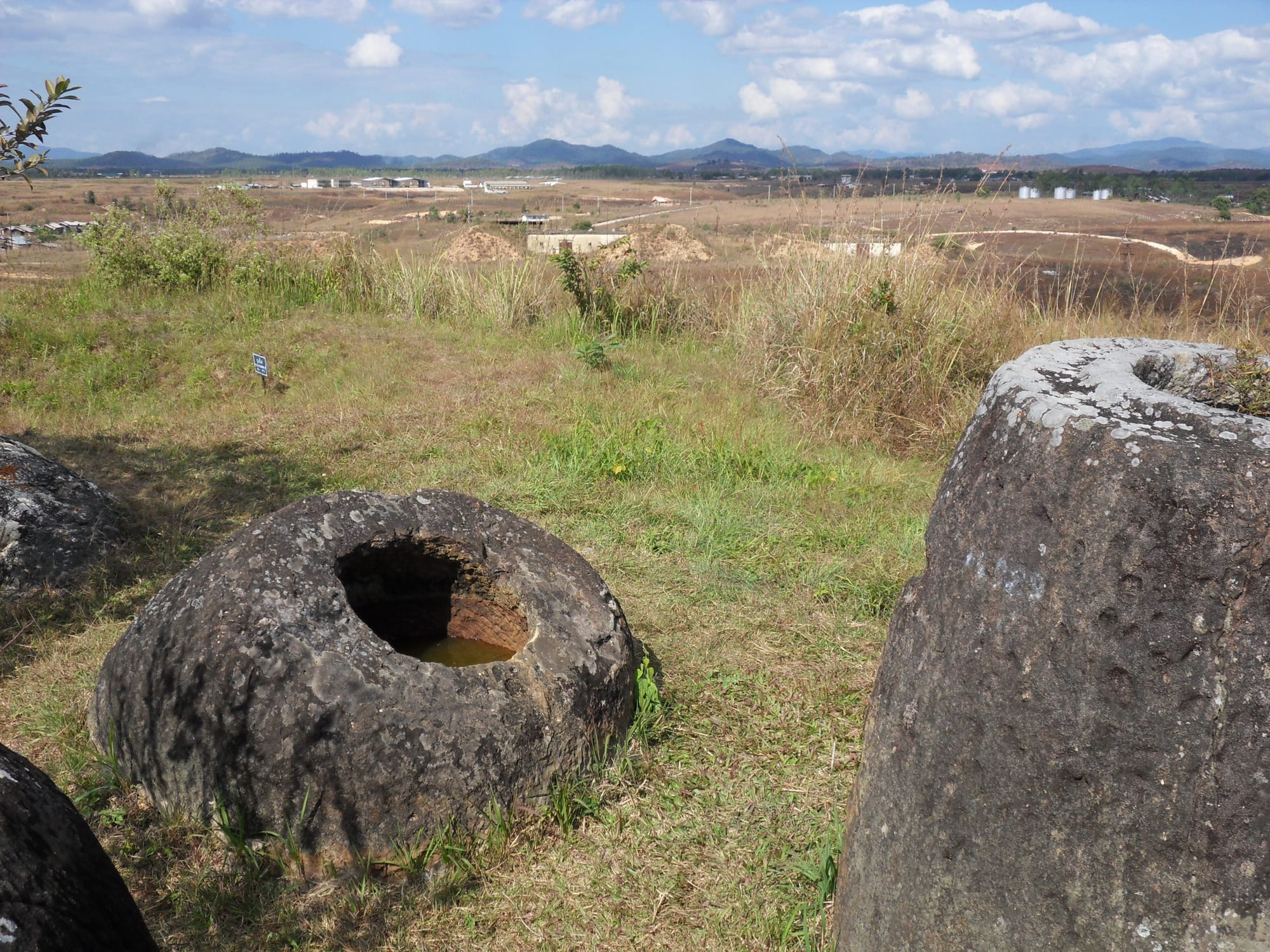 Large weathered stone jars in dry grass, with a wide plain and distant hills behind