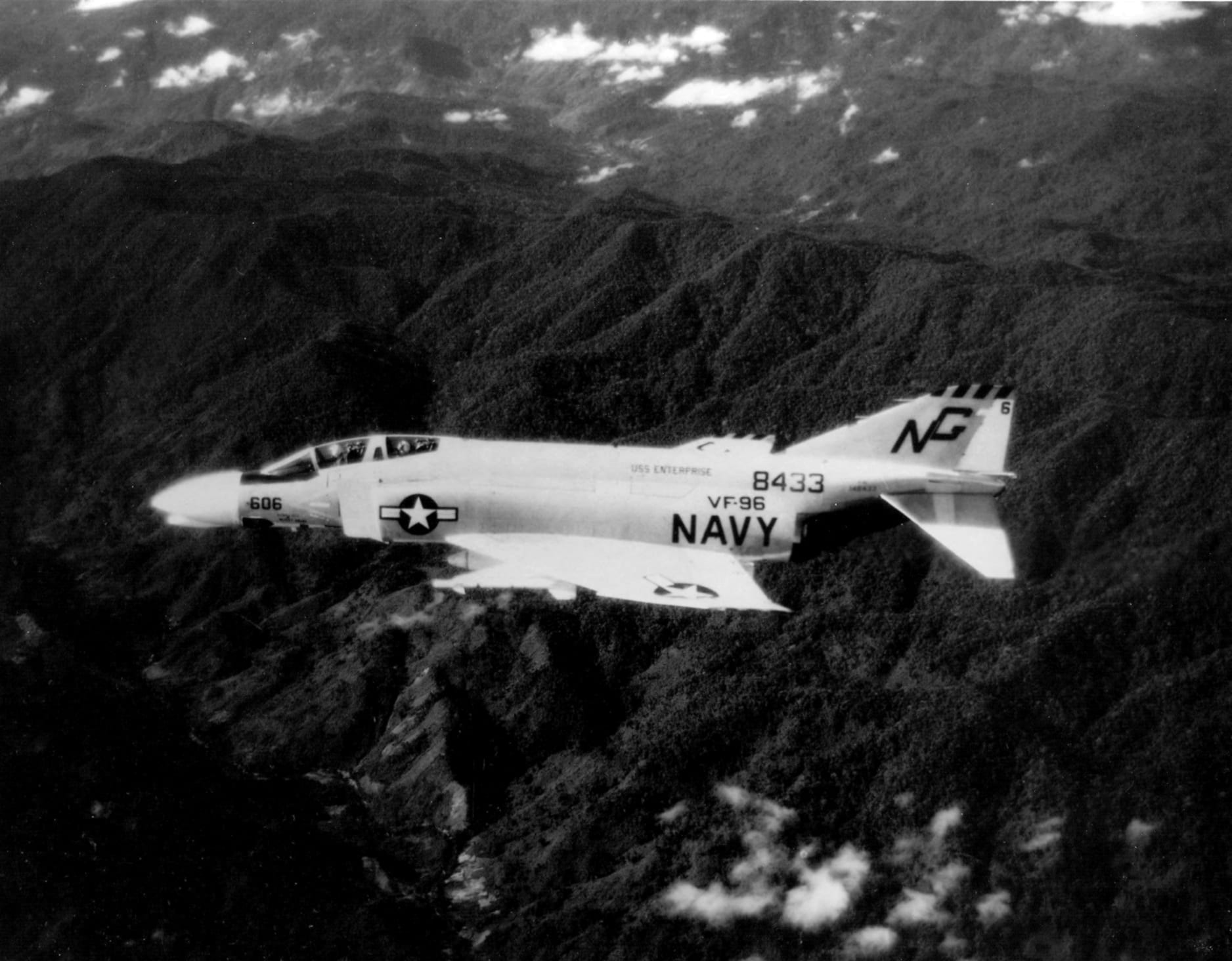A black-and-white photograph of a US Navy jet fighter flying over forested mountain ridges