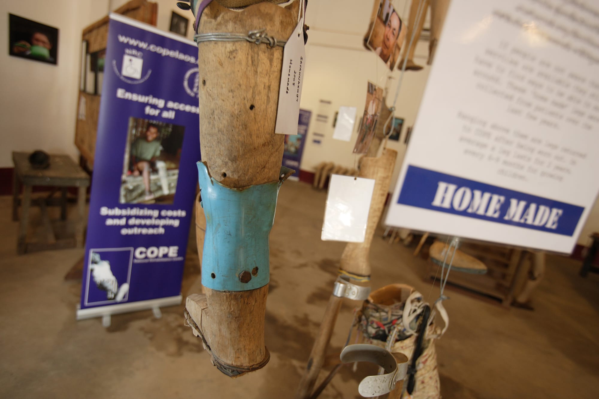 A crude wooden artificial leg with a blue metal brace hanging in an exhibition room, with banners and display boards behind