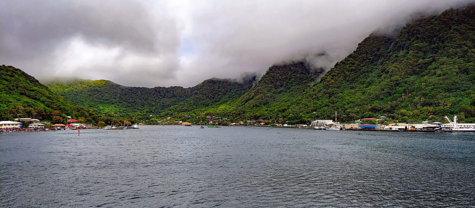 A wide grey harbour ringed by steep jungle-covered mountains with their tops in cloud, and a town strung along the shore