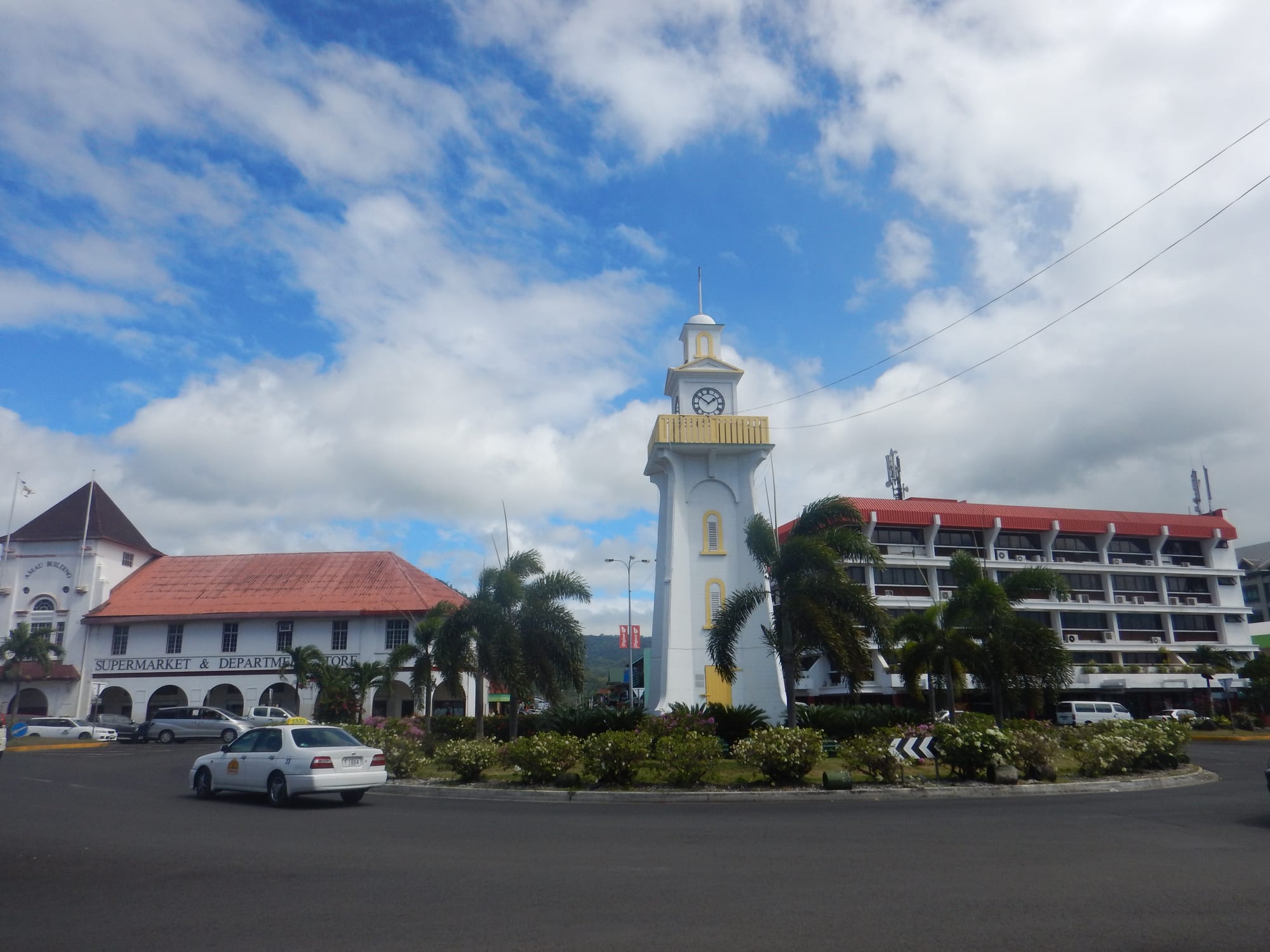 A white clock tower with a yellow balcony on a roundabout lined with palms, between colonial-style and modern buildings