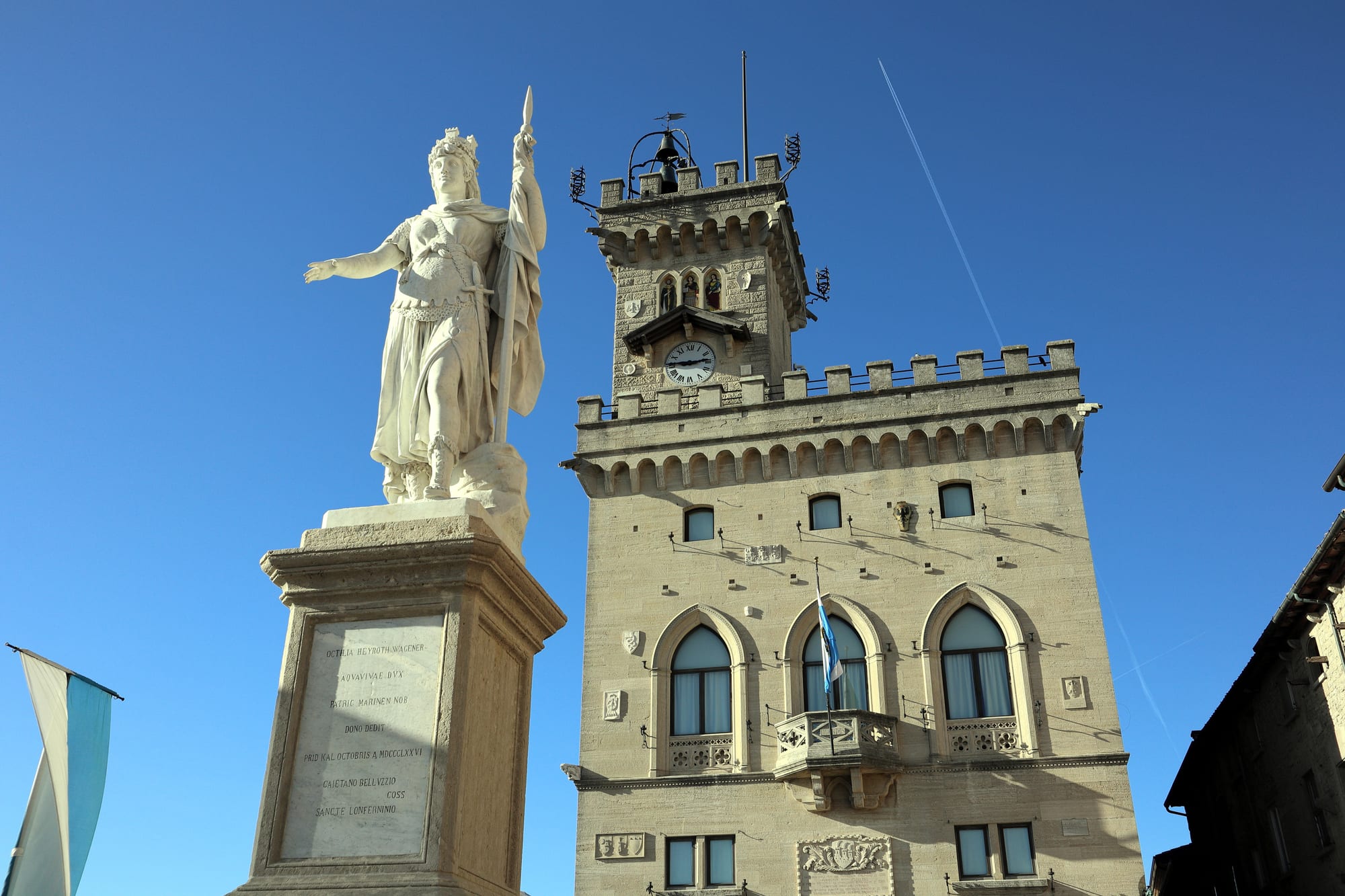 A white marble statue of a crowned woman holding a staff on a tall plinth beside a stone town hall with a battlemented clock tower, under a clear blue sky