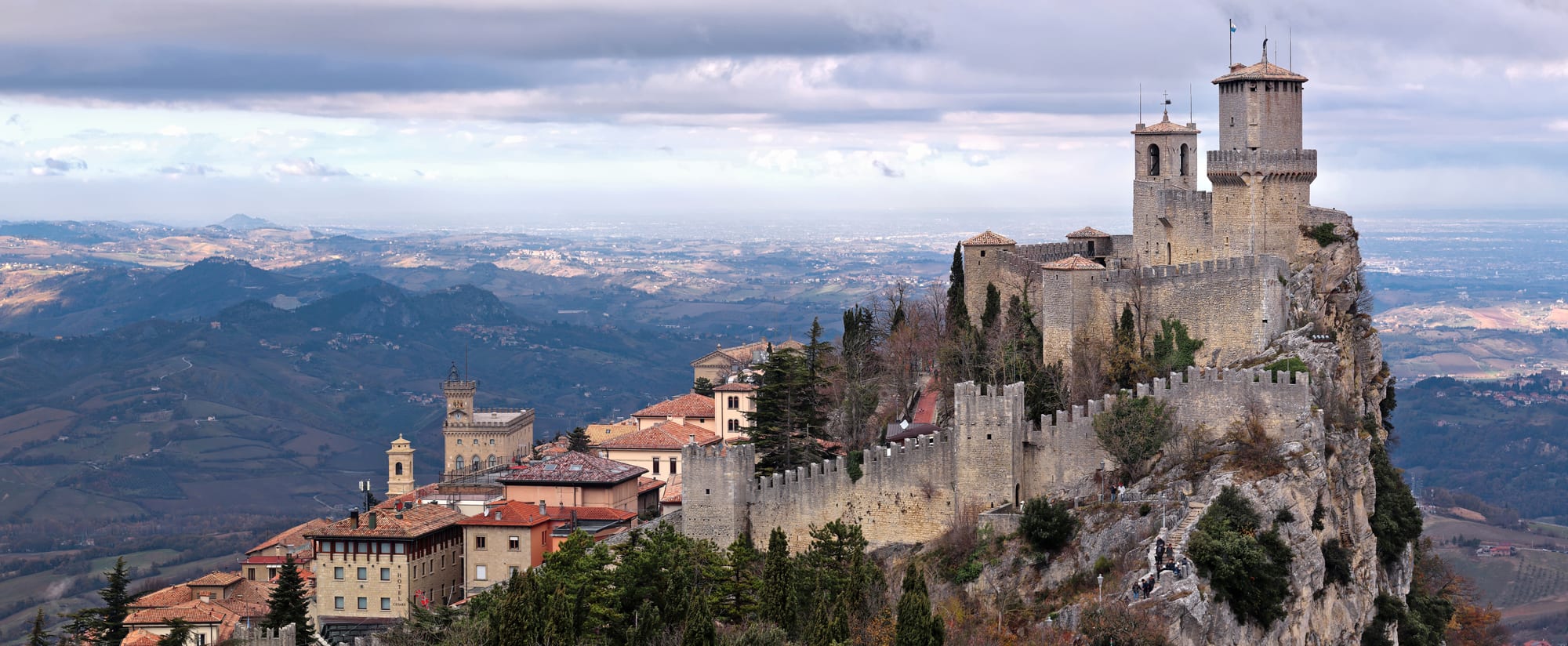A crenellated stone fortress with two square towers on the edge of a rocky cliff, with a red-roofed town below and hills stretching to the horizon