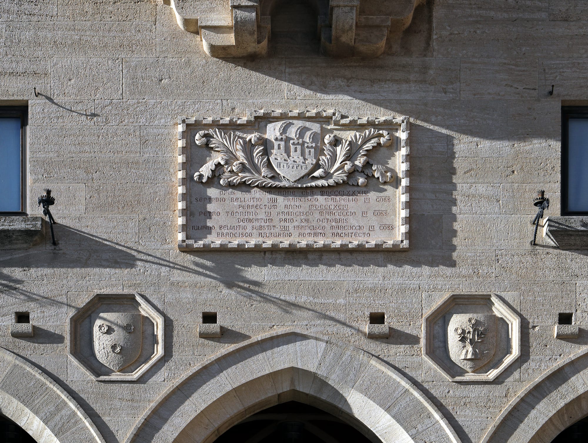 A carved stone plaque with a shield showing three towers on three peaks above the word Libertas, set into a stone facade between two smaller carved shields