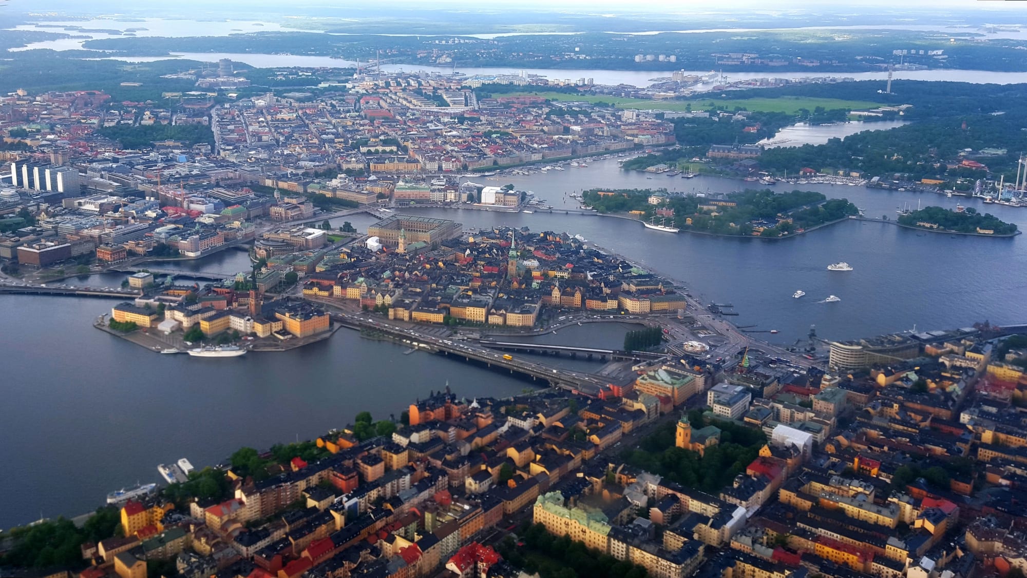 An aerial view of a city spread across several islands, with a dense old town on a central island linked by bridges, wooded islands to the right and a lake stretching away into the distance