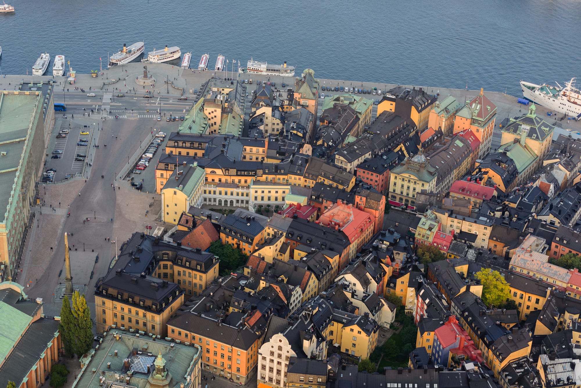 An aerial view of tightly packed old buildings with coloured facades and steep roofs, a wide quay below them and white ferries moored along the waterfront