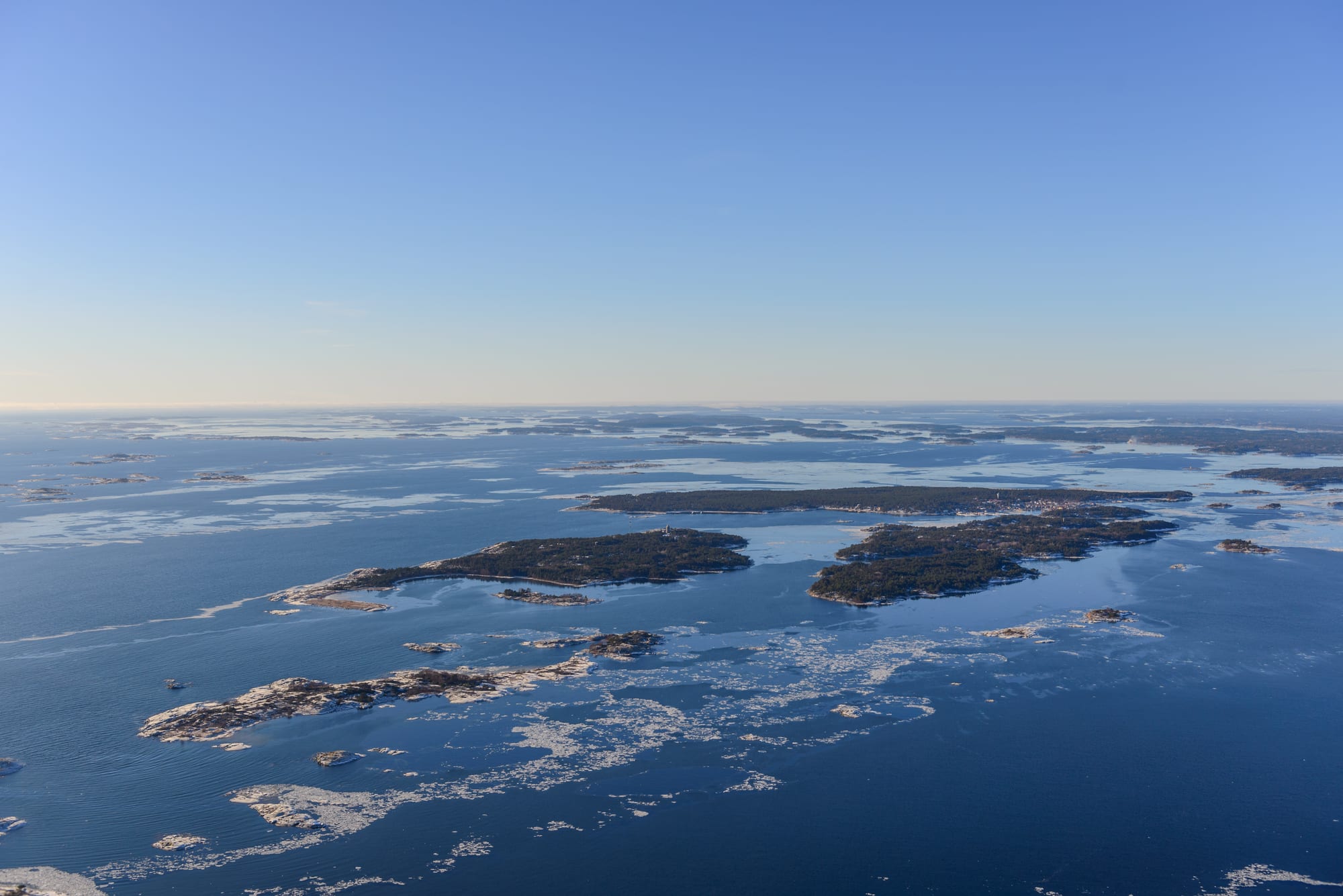 An aerial view of low wooded islands and bare rocky islets scattered across a blue sea patched with winter ice
